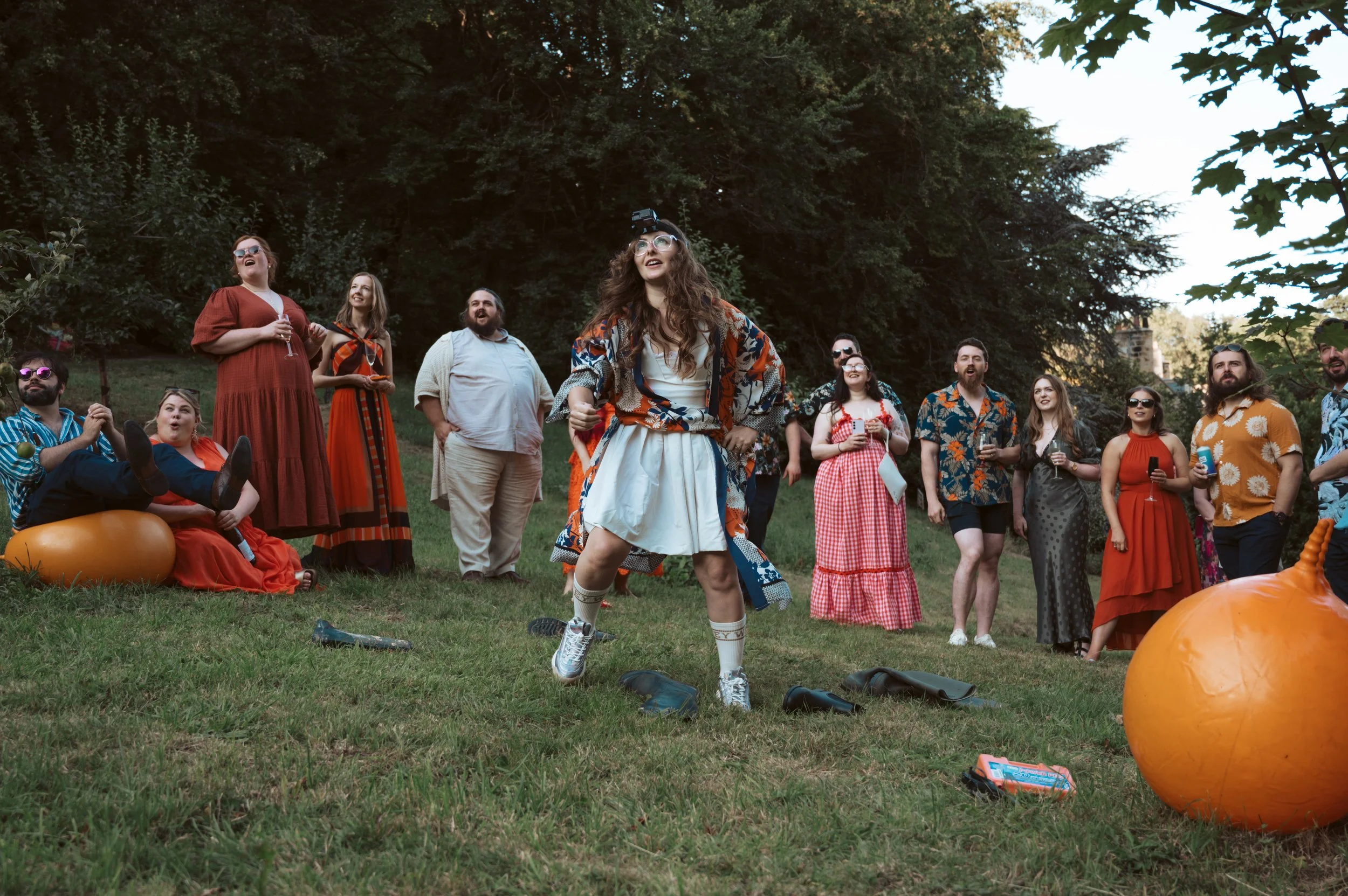 Group of people in colorful clothing standing on grass, watching a woman in a white dress and sneakers with a patterned robe dancing with a humorous expression, surrounded by pumpkins and outdoor scenery.
