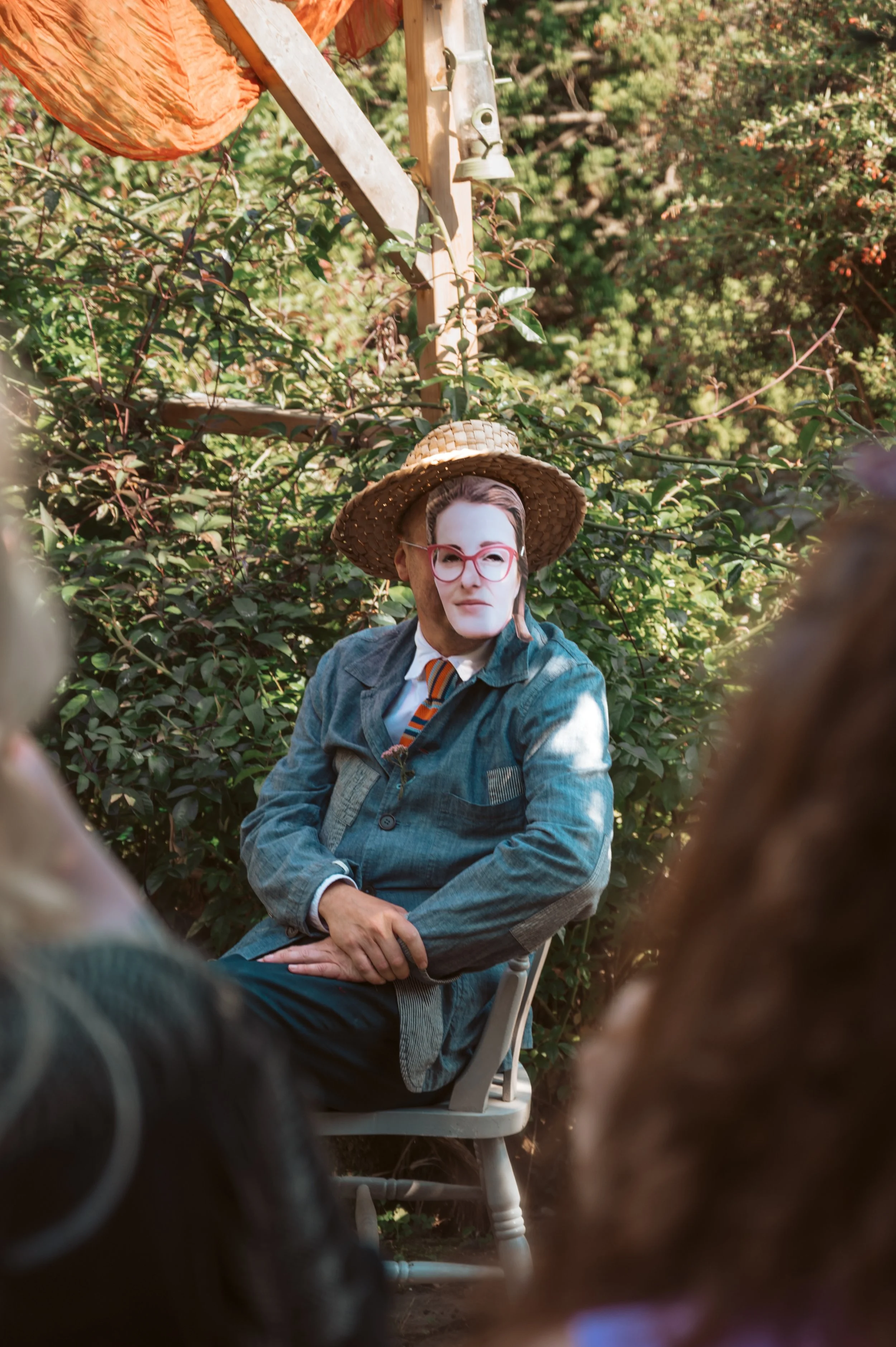 Person wearing a carnival mask, straw hat, glasses, and a vintage-style jacket, sitting outdoors on a wooden chair surrounded by green bushes and trees.
