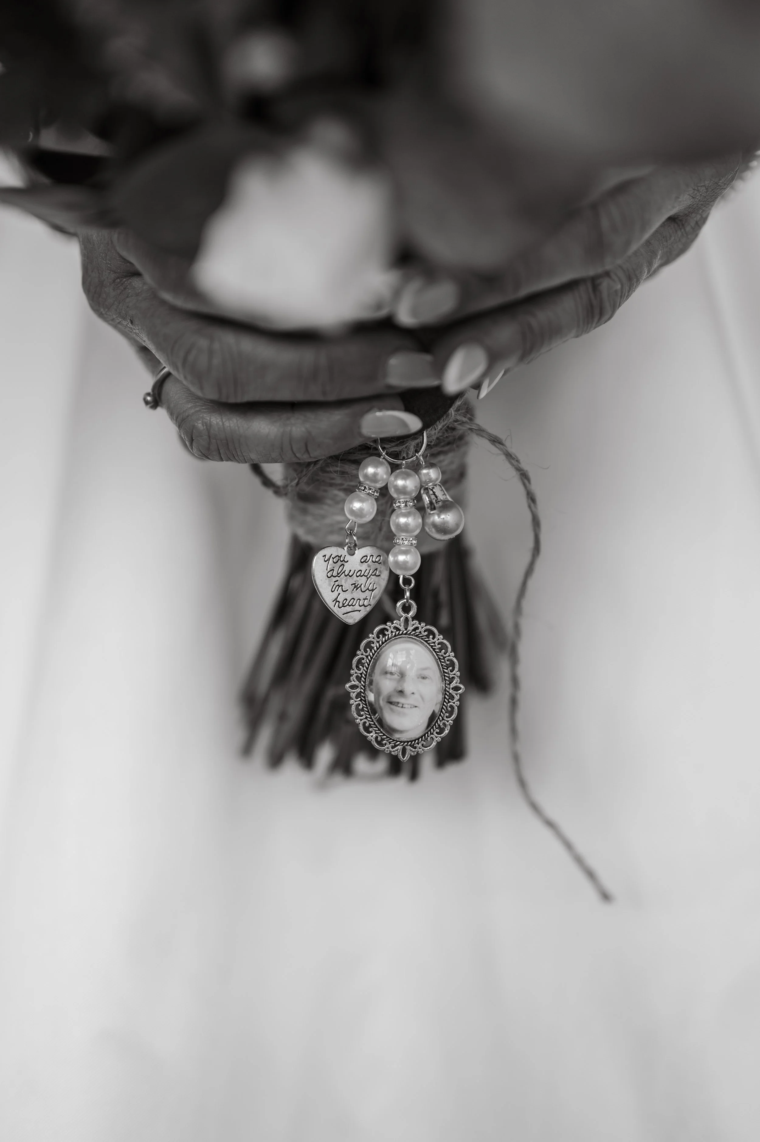 Close-up of a person's hand holding a bouquet of flowers wrapped in dark paper, with a charm bracelet hanging from the wrist featuring a photo of a smiling man, pearl beads, and a heart-shaped pendant with the inscription 'you are always in my heart'