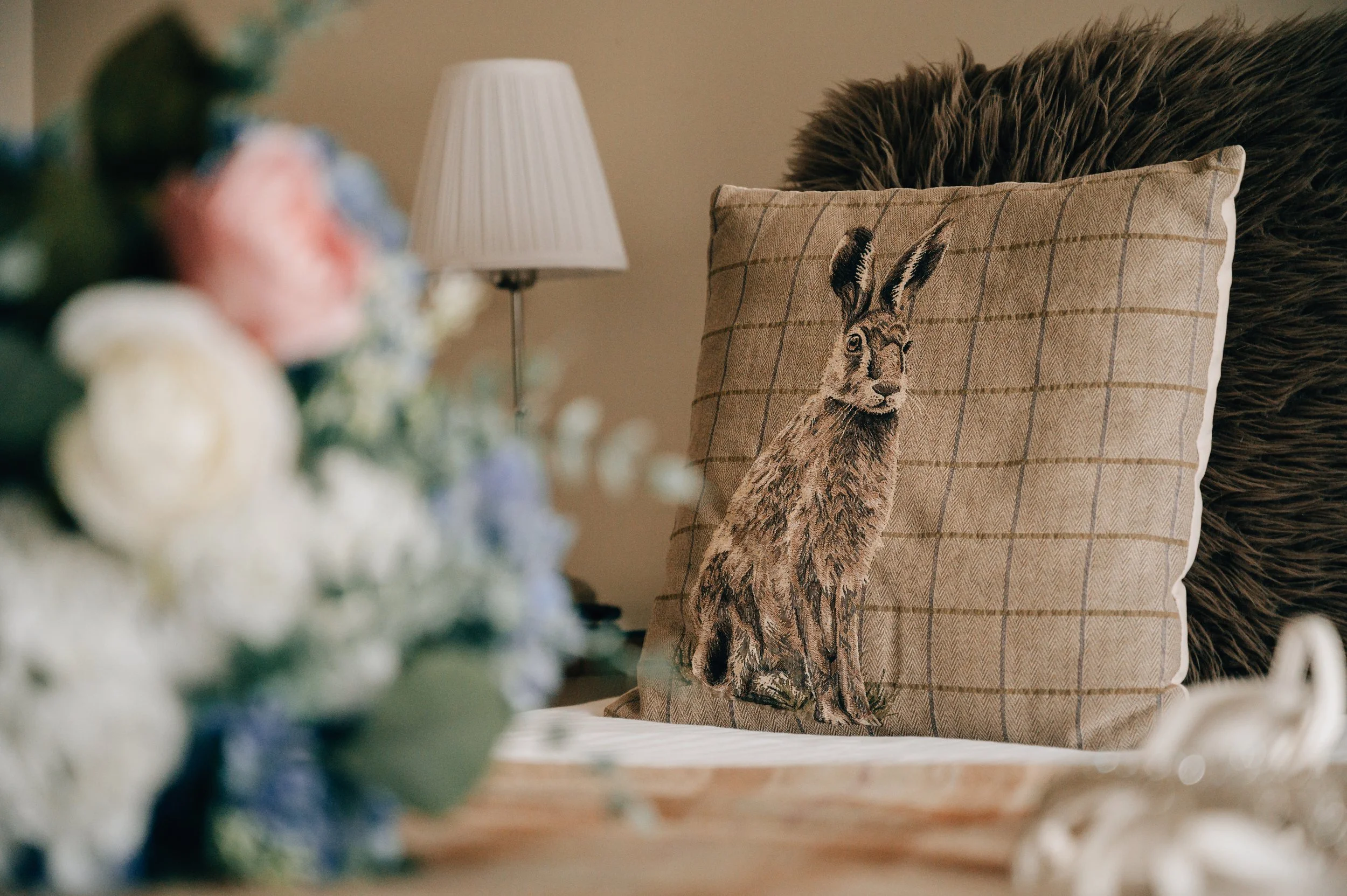 Decorative pillow with a print of a hare sitting on a bed with a fluffy blanket; background includes a blurred bouquet of flowers, a table lamp, and beige walls.