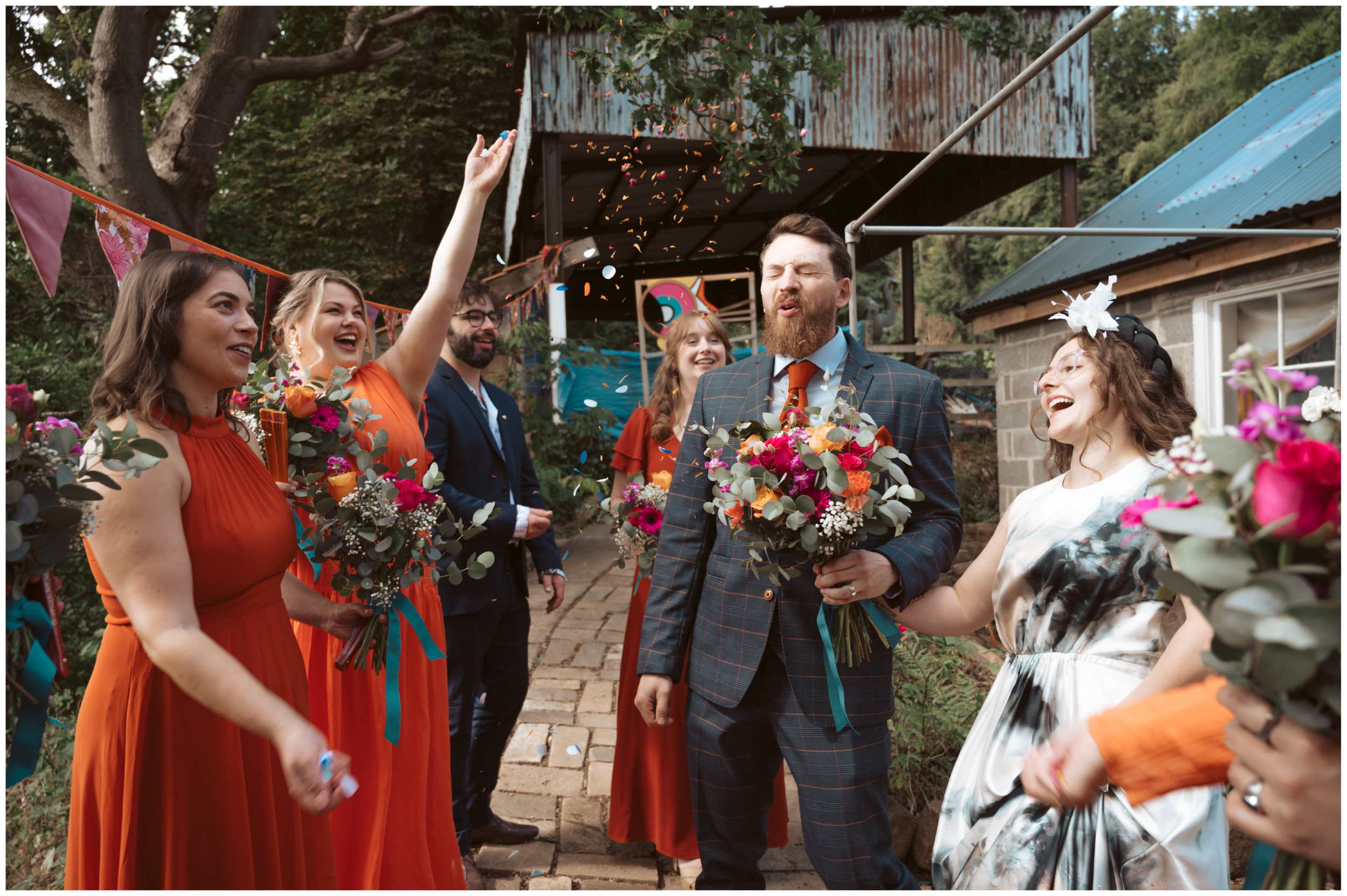 Group of people celebrating outdoors, kissing, laughing, and throwing confetti, with bouquets of flowers in hand, in a garden with trees and a rustic building in the background.
