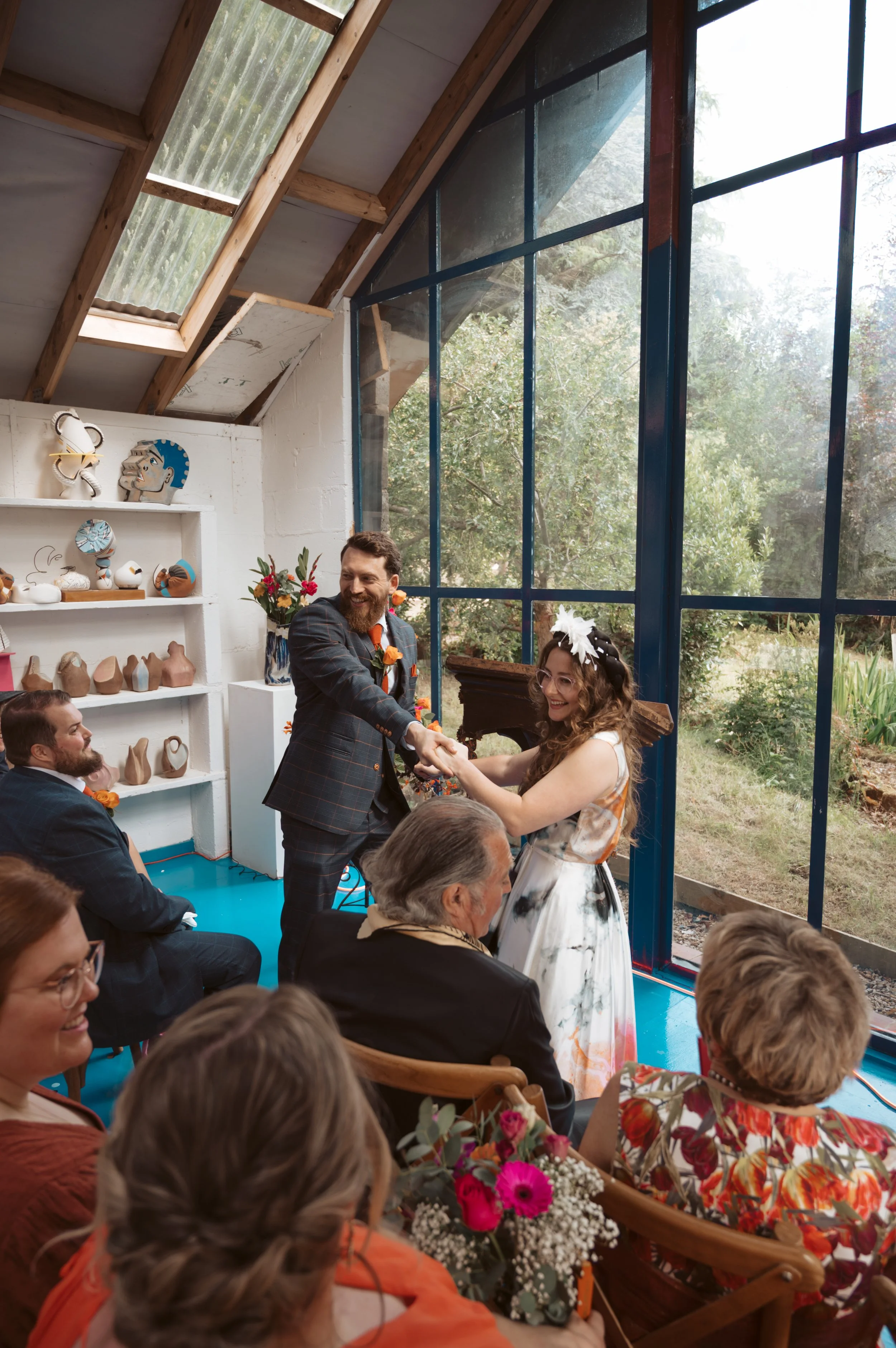 A wedding ceremony with a man and woman holding hands and smiling inside a bright, glass-walled room, surrounded by seated guests, colorful flowers, and decorative pottery on shelves.