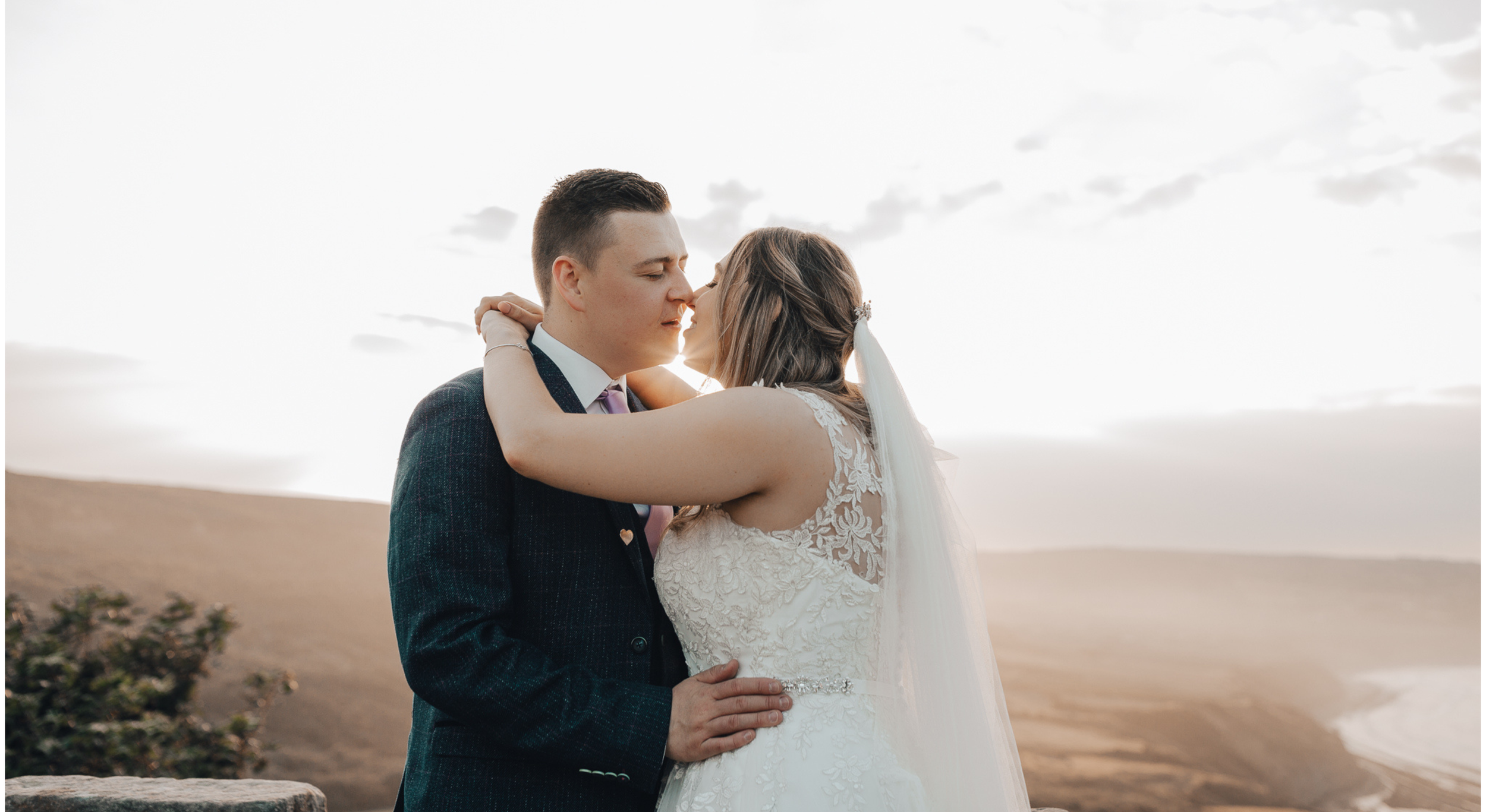 A newlywed couple sharing a romantic moment outdoors at sunset, embracing with eyes closed, overlooking a scenic landscape with rolling hills and a cloudy sky.