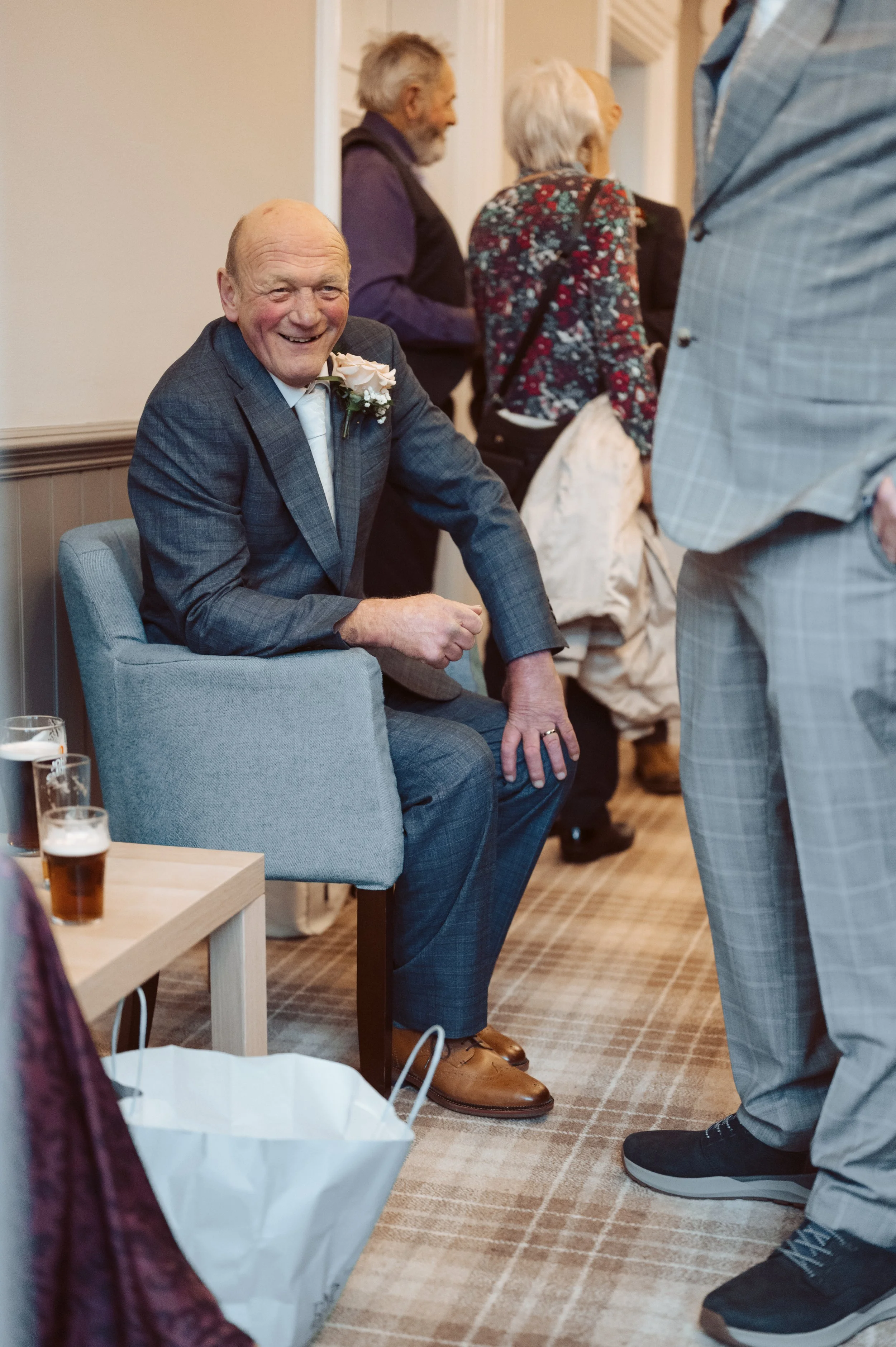 An elderly man in a gray suit with a flower boutonniere sits smiling in a chair at a social gathering, with other older adults in the background and glasses of beer on a table nearby.