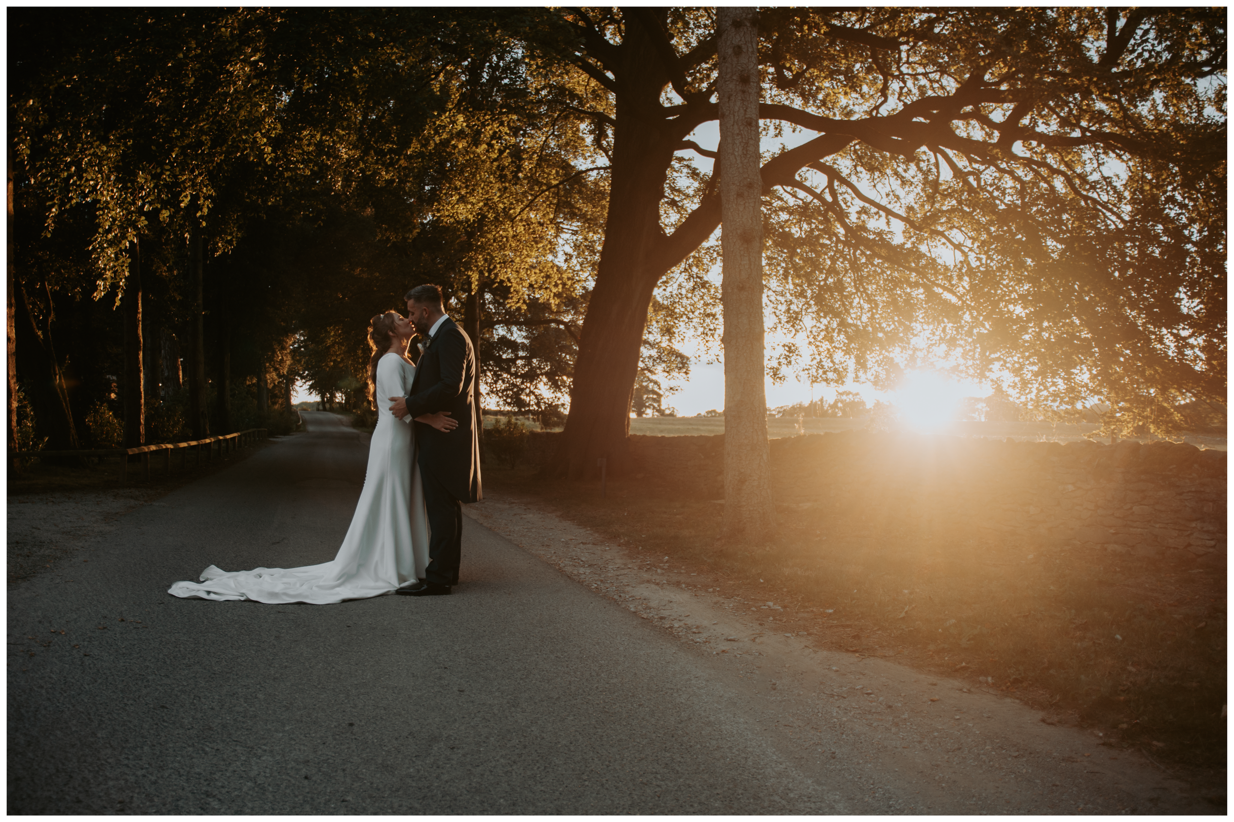 A newlywed couple stands on a country road at sunset, embracing and about to kiss.