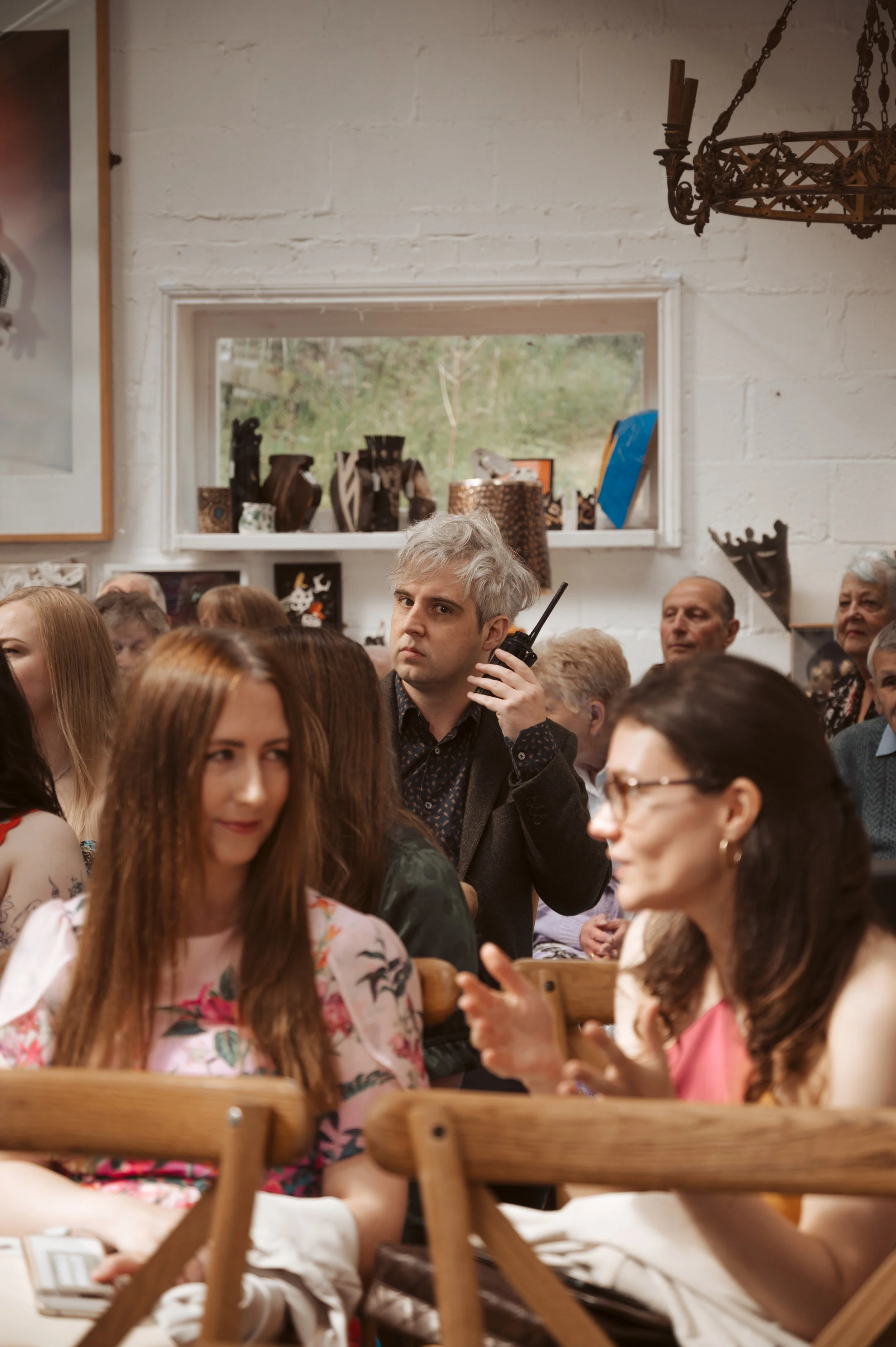 A group of people sitting at tables in a room, with a man holding a walkie-talkie and looking suspiciously at others.