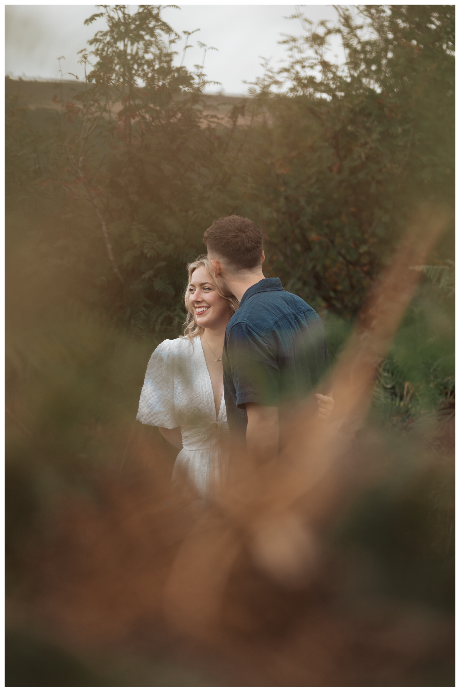 A smiling woman with blonde hair in a white dress standing close to a man with short brown hair in a dark shirt, outdoors surrounded by trees and foliage, seen through blurred branches.