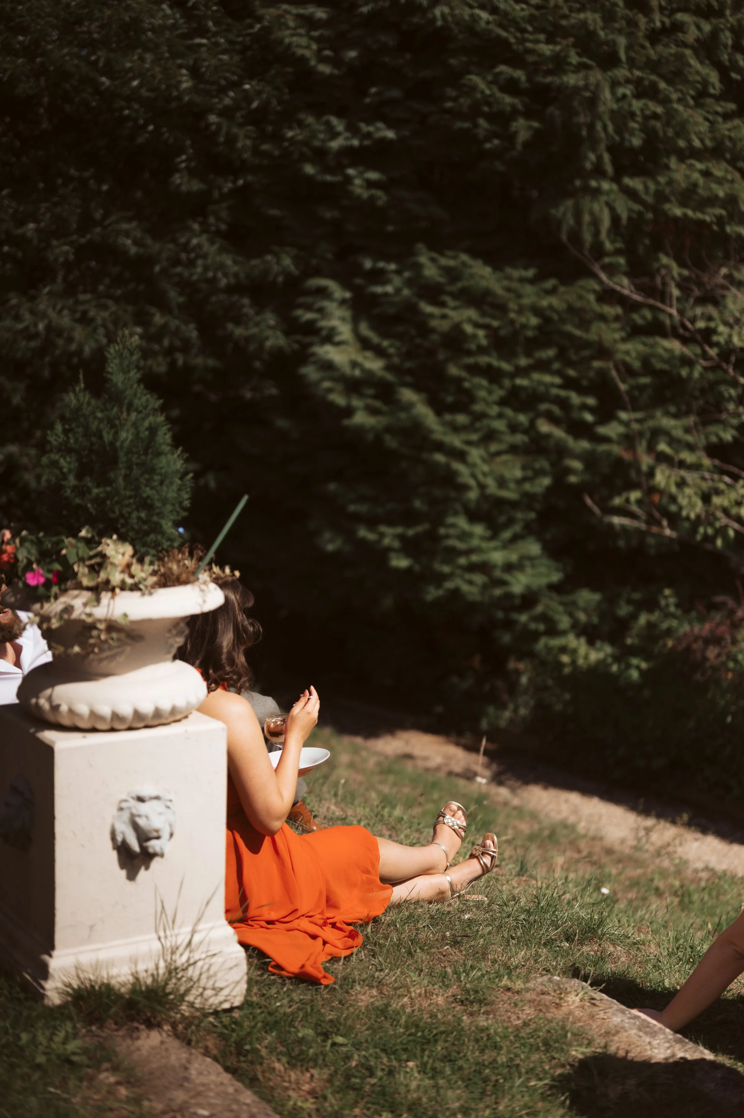 A woman in an orange dress sat on the grass in a garden, holding a glass, with trees in the background and a decorative stone planter nearby.