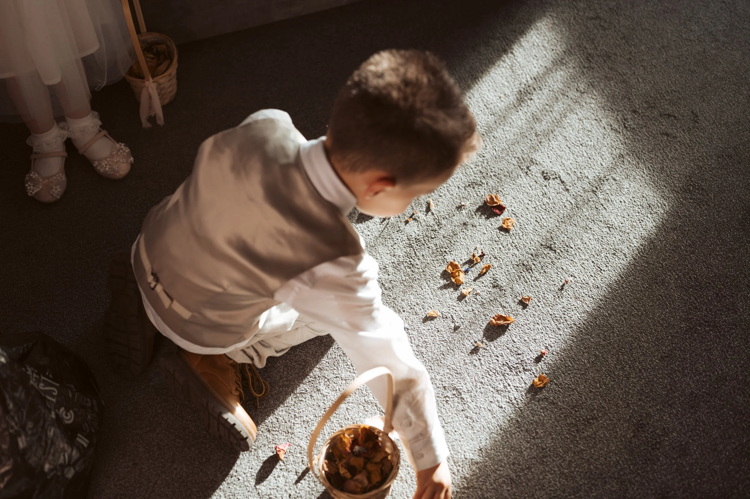 A young boy in a white shirt and beige vest, sitting on the carpet, reaches into a basket filled with dried leaves, while leaves are scattered on the carpet in front of him. Sunlight streams through a window, casting shadows on the floor.