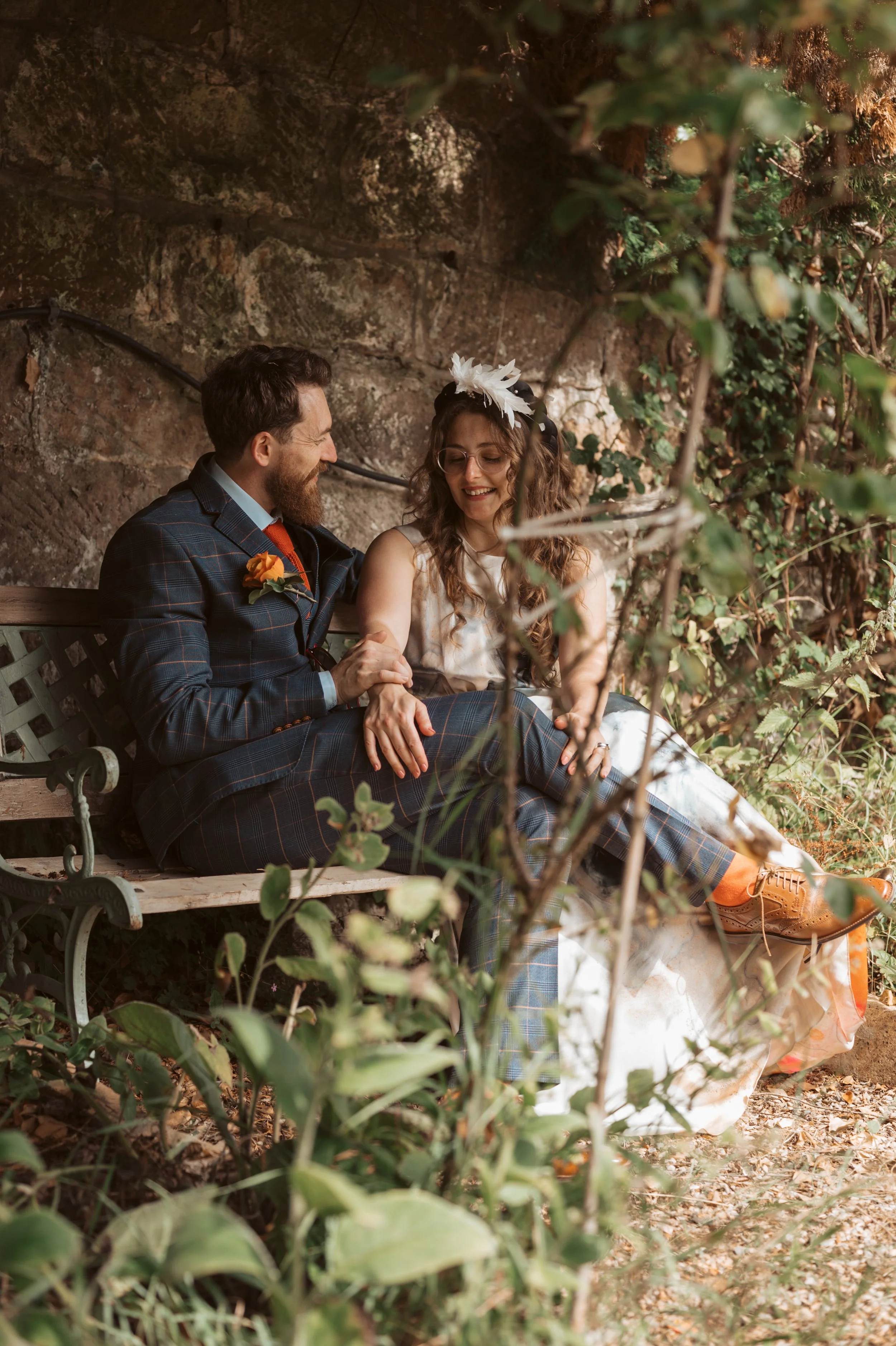 A joyful couple sitting on a park bench, holding hands and smiling at each other. The man is dressed in a plaid suit with a boutonniere, and the woman is wearing a white dress with a floral headpiece. They are surrounded by greenery and a stone wall.