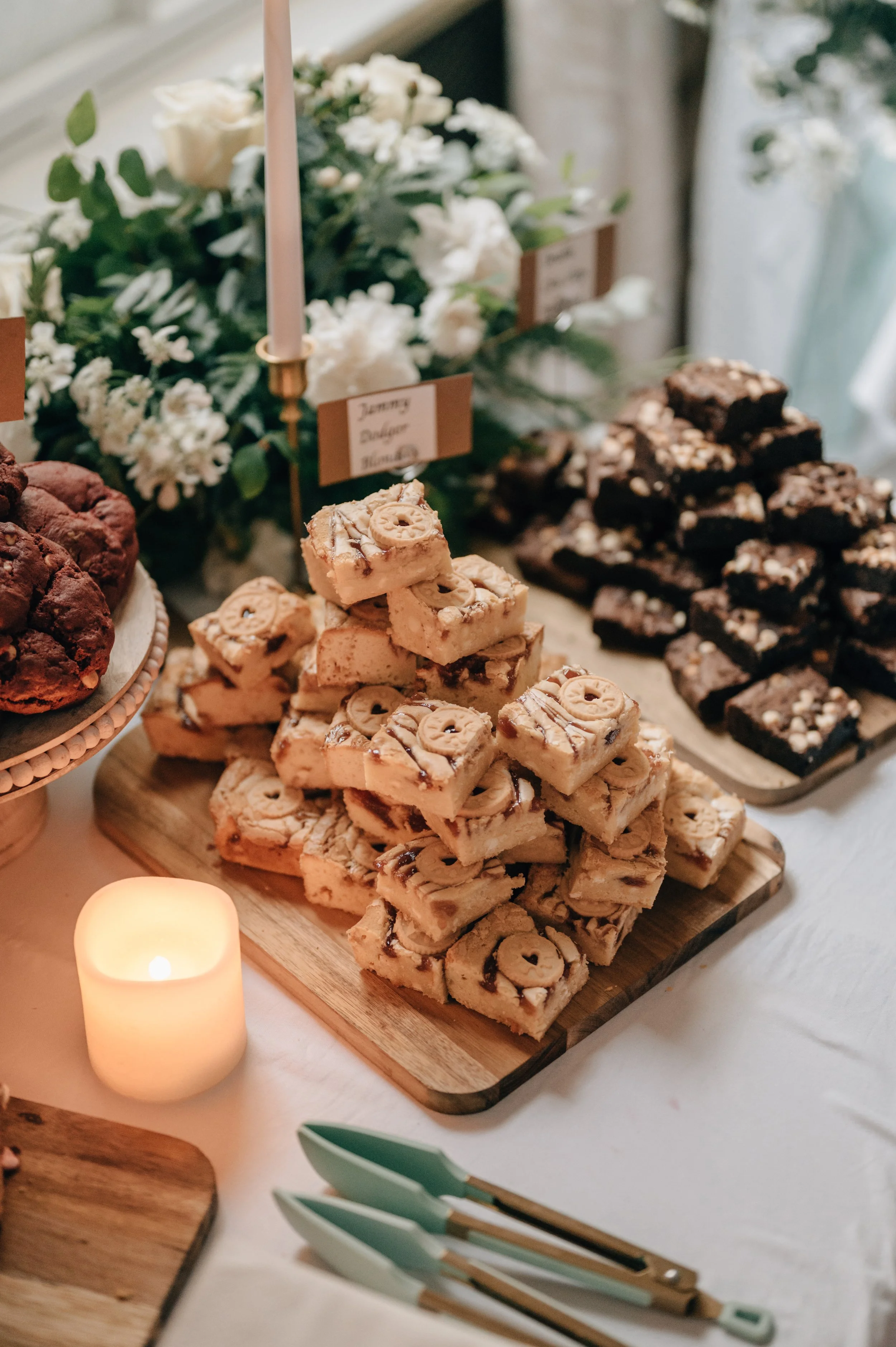 A dessert table featuring a pyramid of cookie bars, a tray of brownies, a plate of chocolate cookies, a lit candle, and a small bouquet of white flowers in the background.