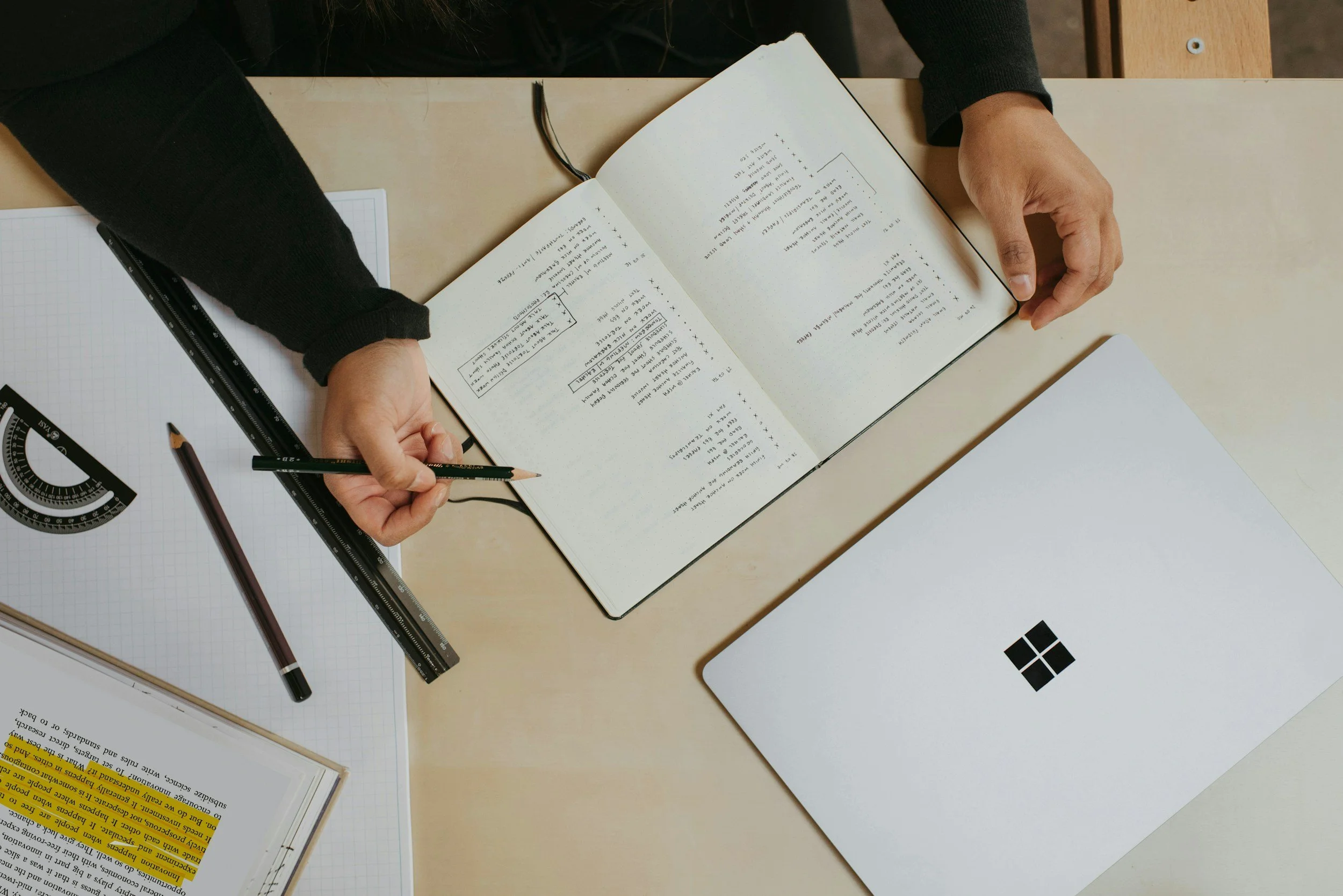 Overhead view of a person holding an open notebook filled with handwritten notes, surrounded by office supplies including a graph paper notebook, a pen, a ruler, and papers on a light-colored desk.