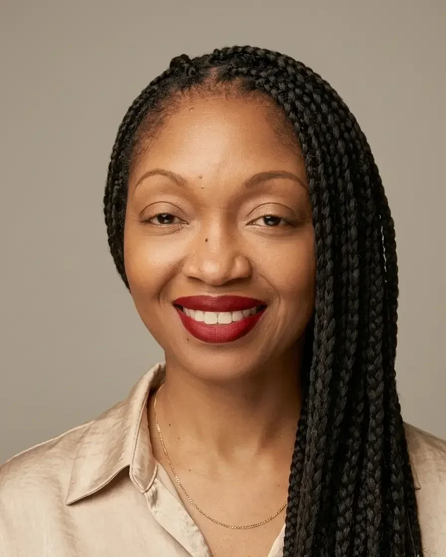 Une femme aux longs cheveux tressés, souriante, portant du rouge à lèvres rouge, une chemise en satin beige et un délicat collier en or, sur un fond neutre.