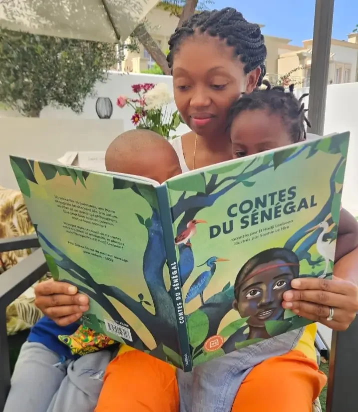 Ingrid Francisque, founder of Afrobudgetingirl, reading a colorful children's book titled 'Contes du Sénégal' to her children, outdoors on a patio with plants and trees in the background.