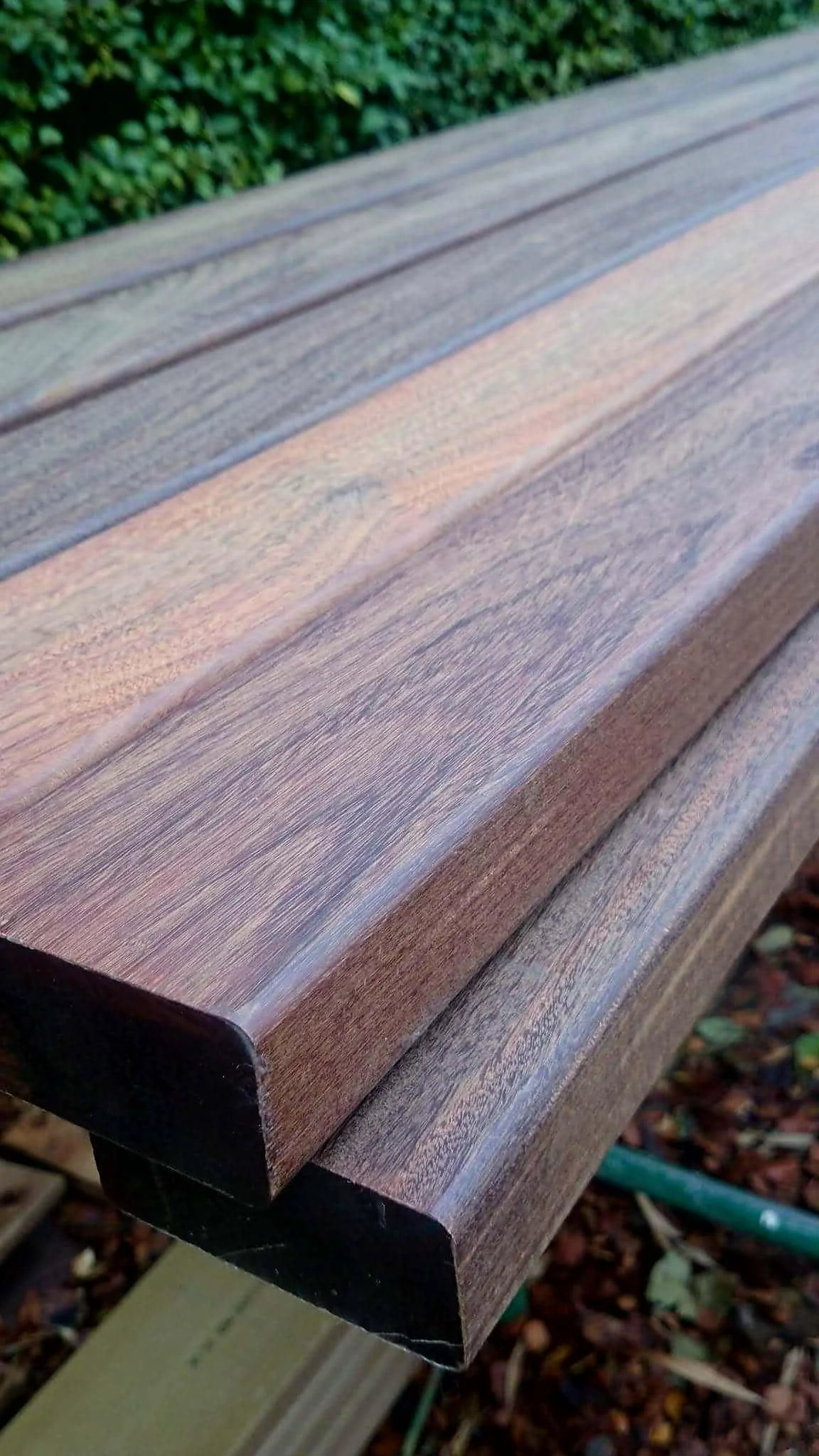 Close-up photo of a wooden bench with a dark edge, outdoors near green bushes and red mulch ground.