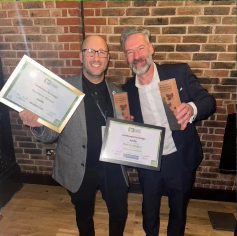 Two men standing in front of a brick wall, smiling and holding awards and certificates. One is wearing glasses and a gray jacket, the other with gray hair and a beard, wearing a dark suit.