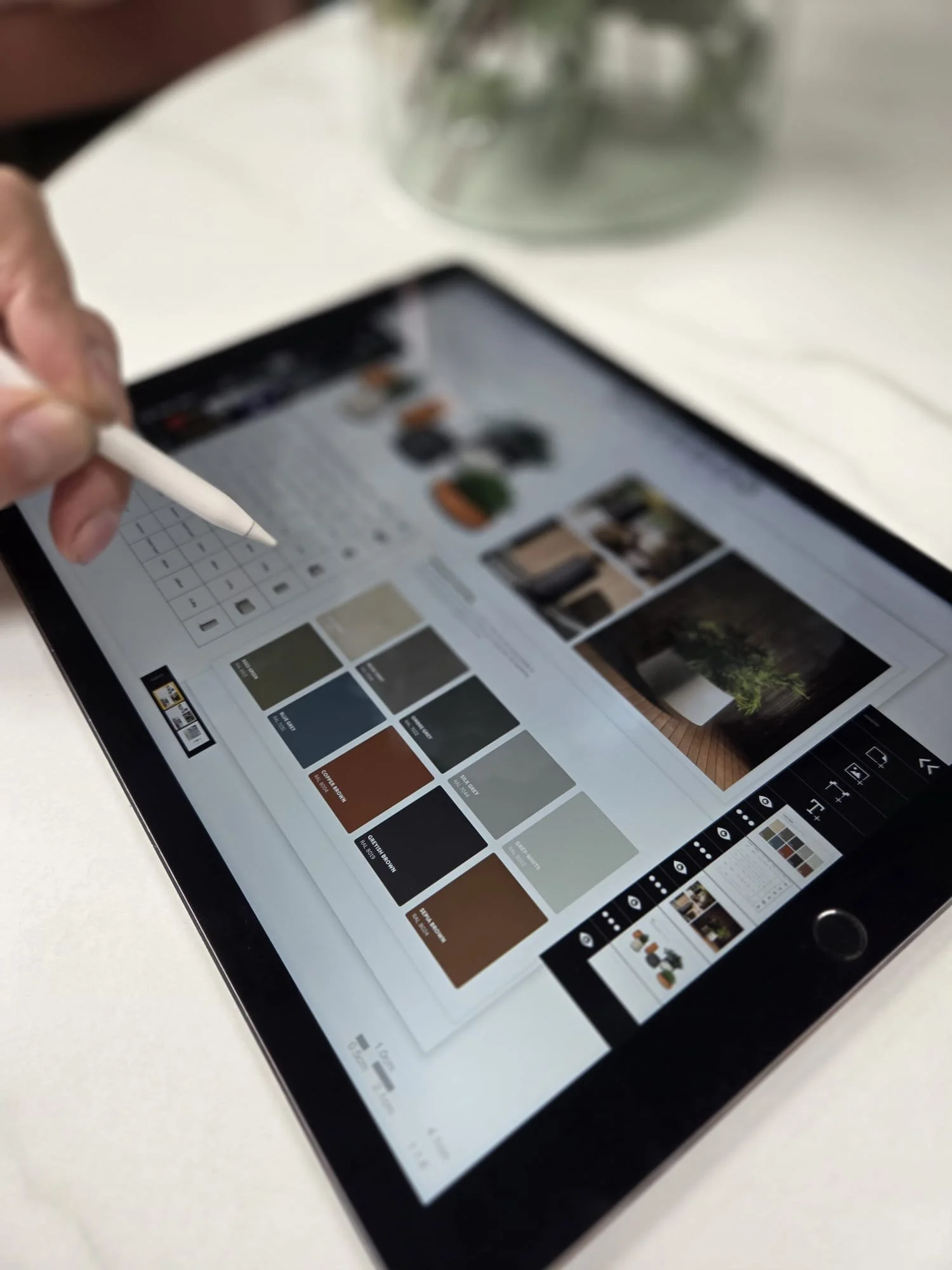 A person's hand holding a stylus drawing on a tablet displaying a design or mood board with color swatches and images, on a white table.