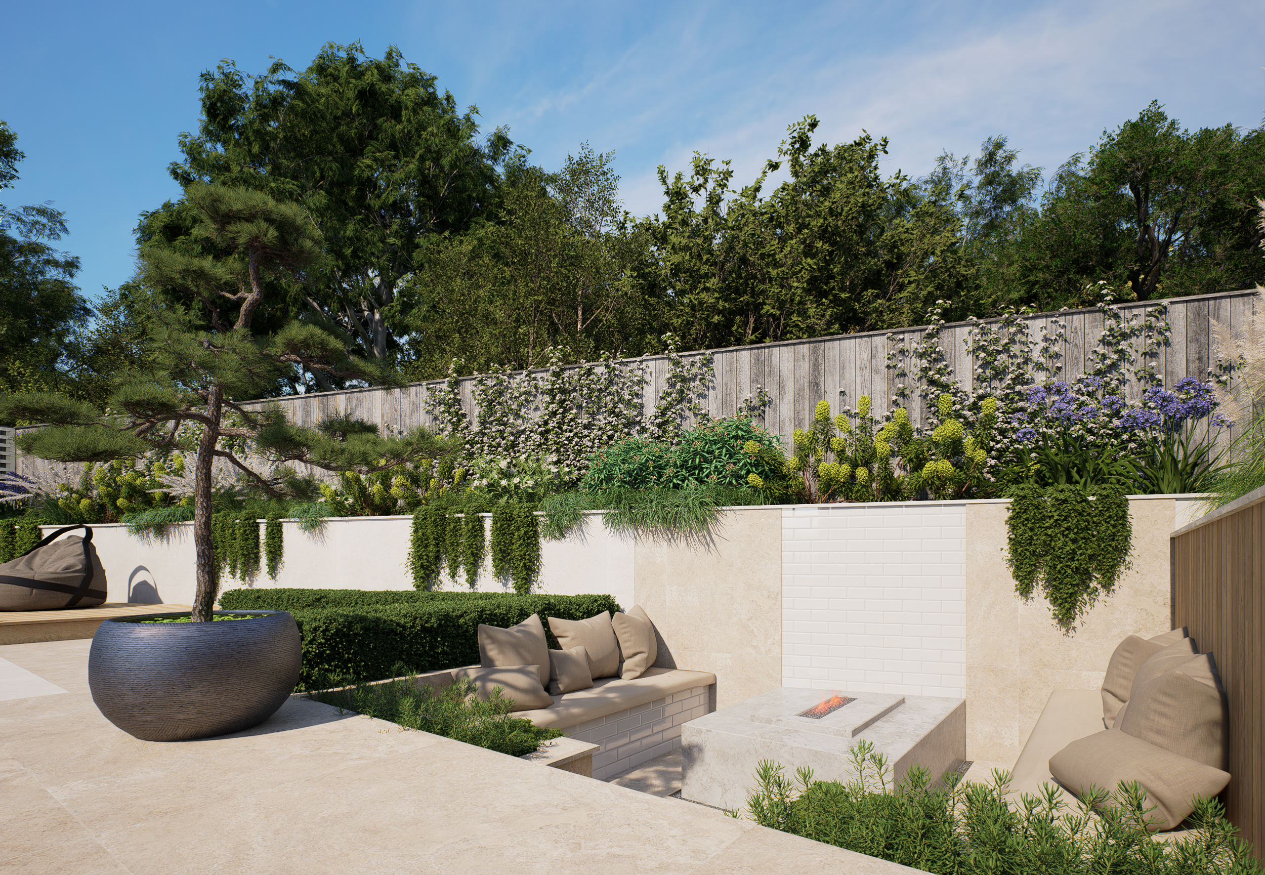 A modern backyard patio featuring beige outdoor furniture with pillows, a large black planter with a small tree, lush green plants along the wall, a small built-in fire pit, and a backdrop of a wooden fence covered with flowering vines and tall trees