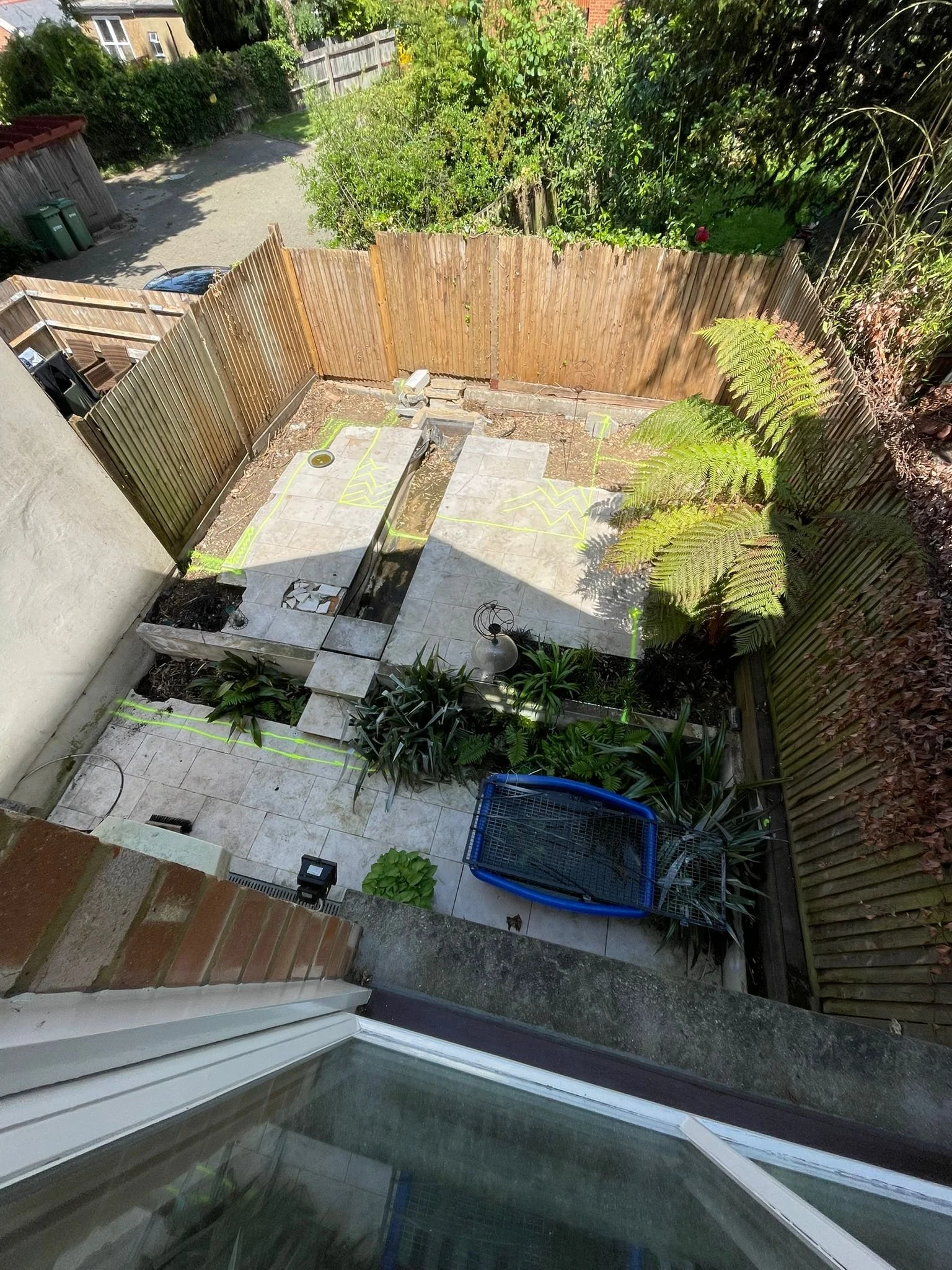Backyard with partially paved area, plants, construction markings, and a blue shopping cart or garden cart. Wooden fence surrounds the yard, with trees and greenery beyond.