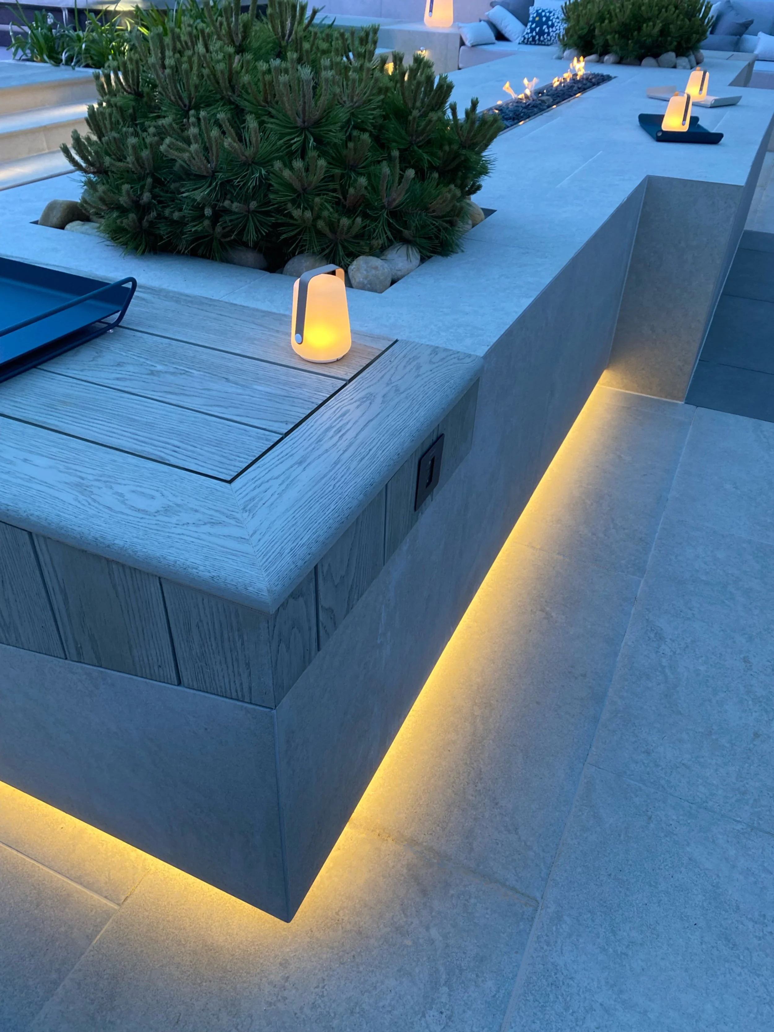 Outdoor modern patio with porcelain and composite decor, illuminated with LED strip lights. Contains a concrete planter with a green bush, small lanterns providing warm light, and a blue tray on the wooden surface.