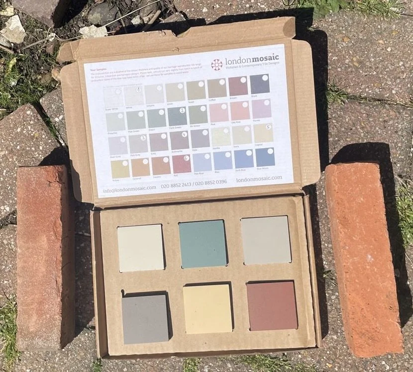 Open cardboard box containing six paint color samples arranged in a grid, with a color chart and contact information on the inside of the lid, placed on brick and concrete ground outdoors.