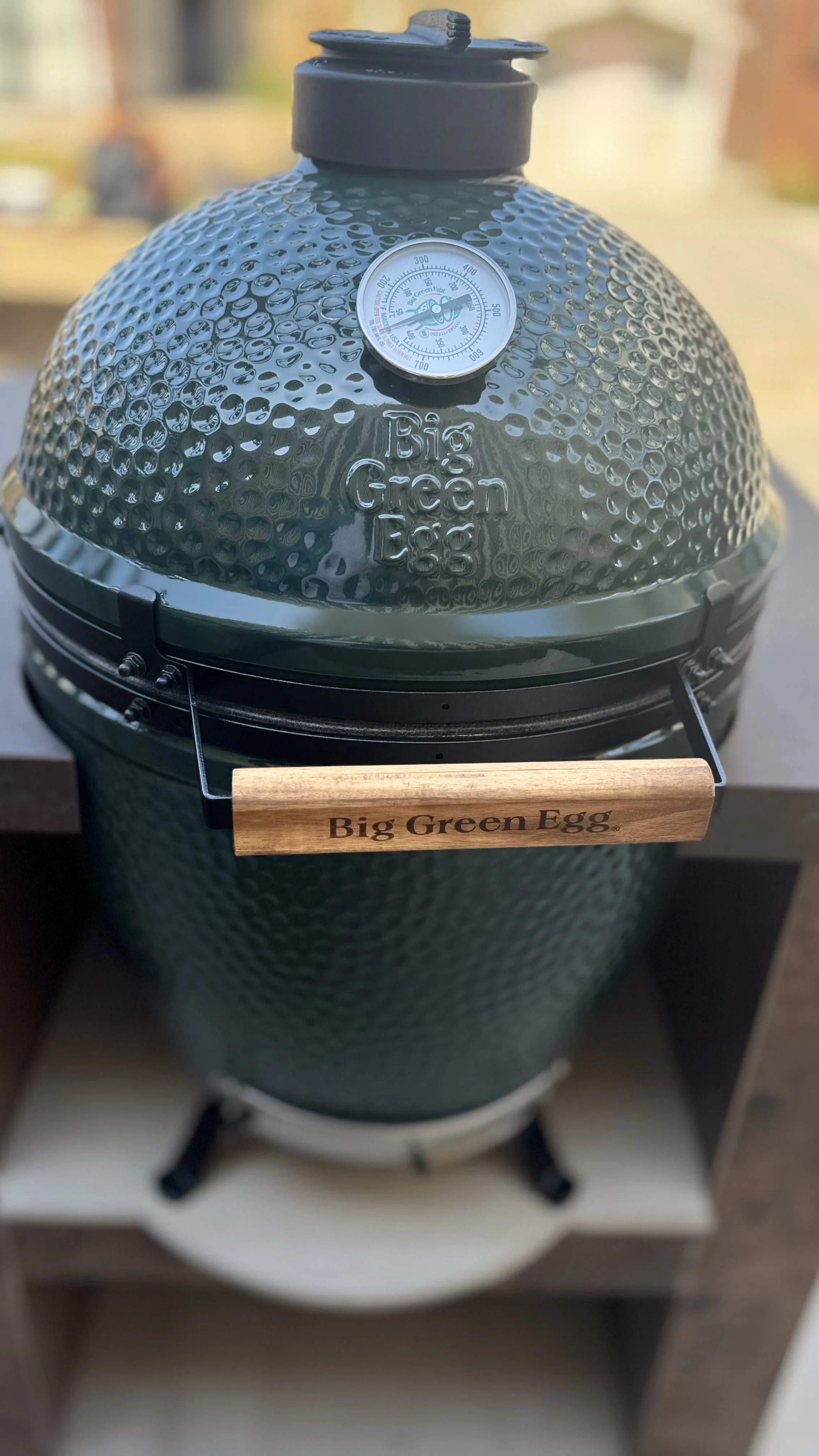 A green Big Green Egg barbecue grill with a textured surface, thermometer on the lid, and wooden handle with the brand name.