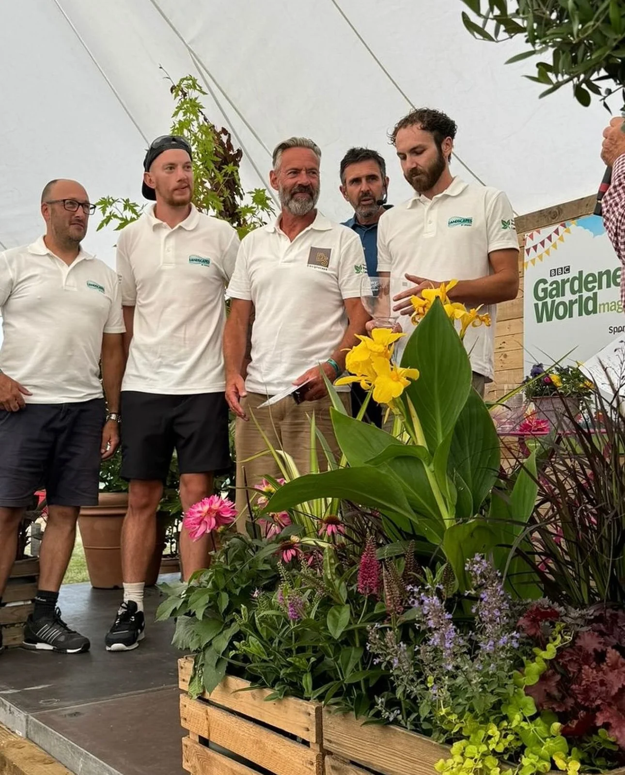 Group of men at a gardening event, some wearing white polo shirts, standing behind a display of vibrant flowers, with a BBC Gardening World magazine banner in the background.