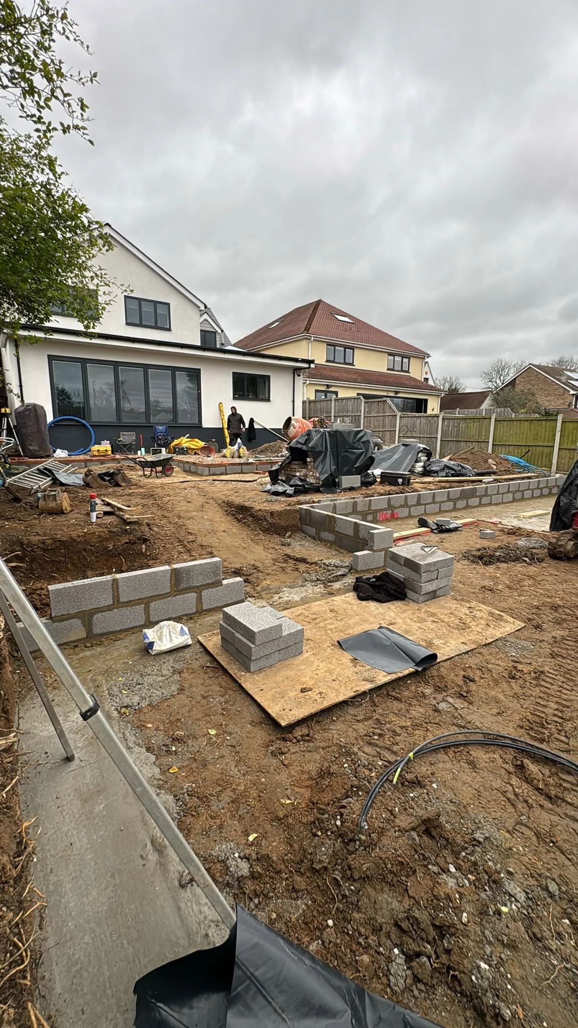 Backyard construction site with masonry work underway, showing brick walls, construction tools, and materials under cloudy skies.