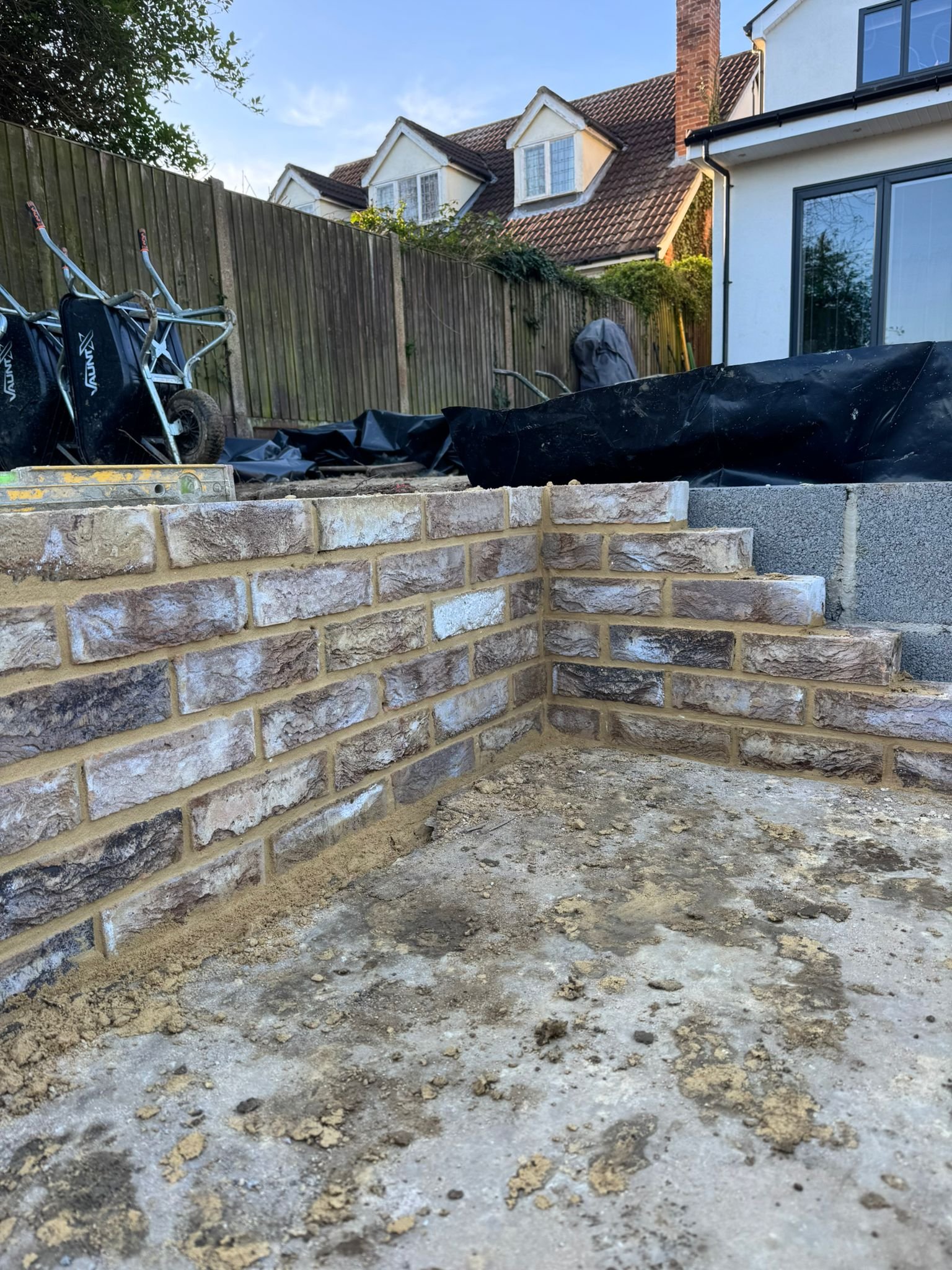 Backyard with partially built brick retaining wall, construction materials, wheelbarrows, and houses in the background.