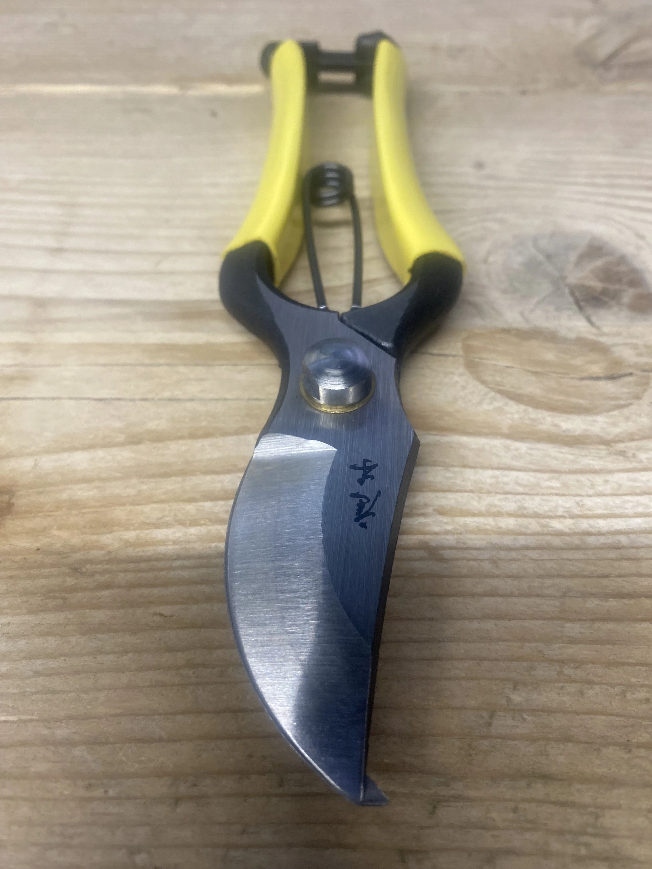 A pair of yellow-handled pruning shears with a curved metal blade resting on a wooden surface.