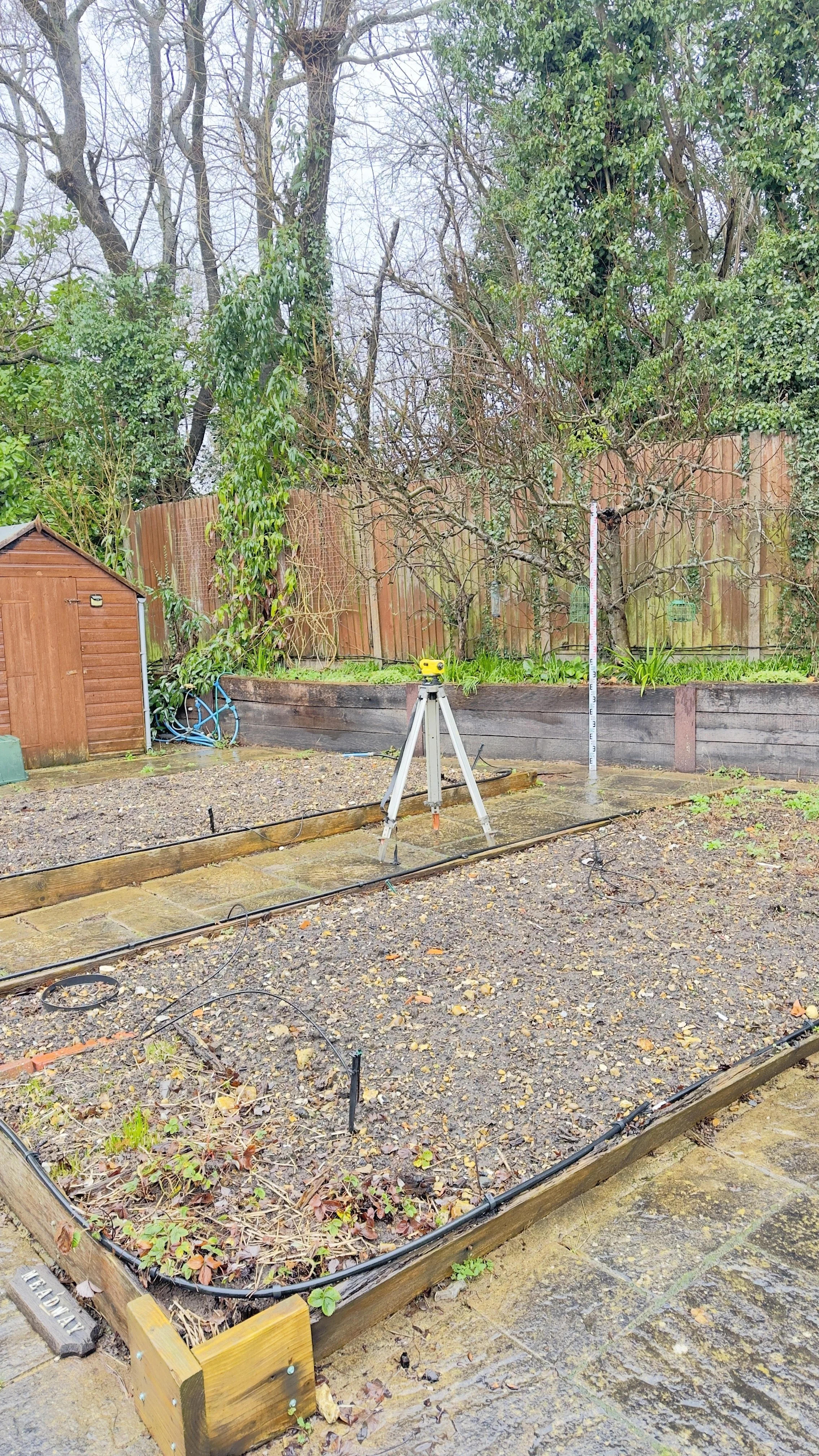 A garden area under construction with a wooden frame around a planting bed, a tripod-mounted laser level in the center, a measuring pole, and wires on wet ground, with trees and a wooden fence in the background.