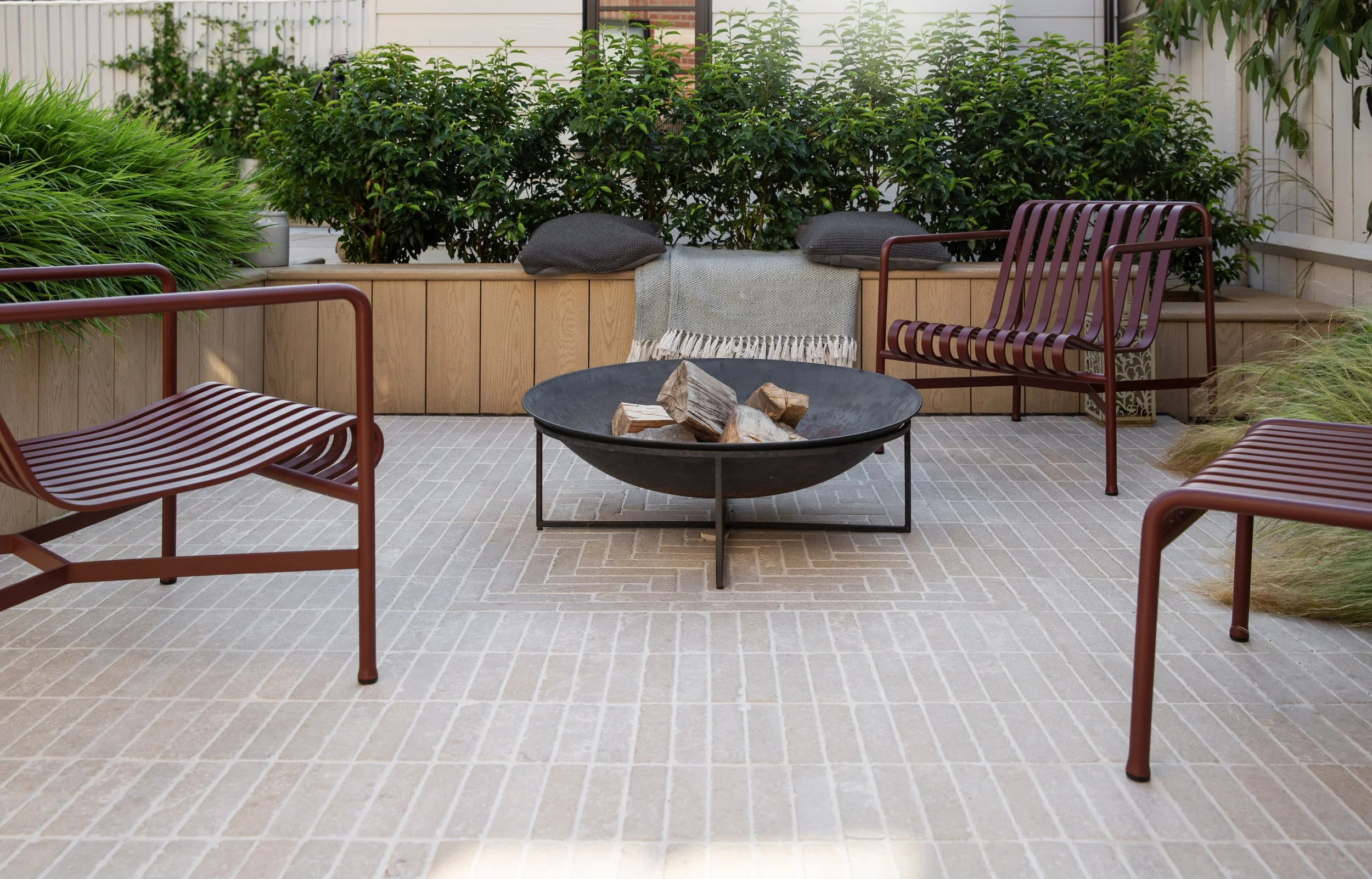 An outdoor patio with a stone-tile floor, surrounded by green plants and shrubs, featuring a black fire pit with wooden firewood, two red metal chairs, and a wooden decorative bench with cushions and a throw blanket on it.