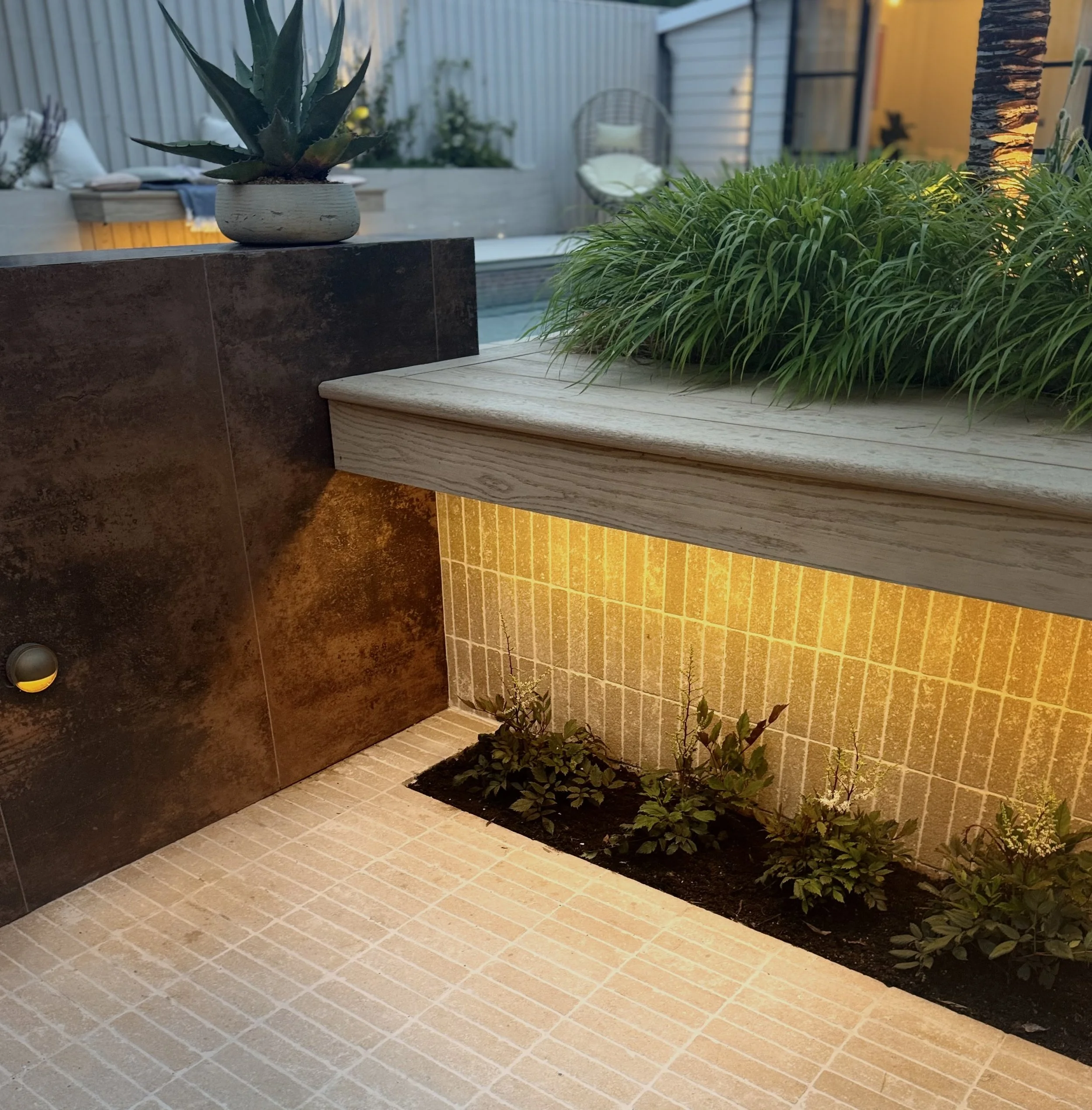 Close-up of a patio corner with potted succulent, a planter with green grass, and a small garden bed with small plants, illuminated by warm lighting.
