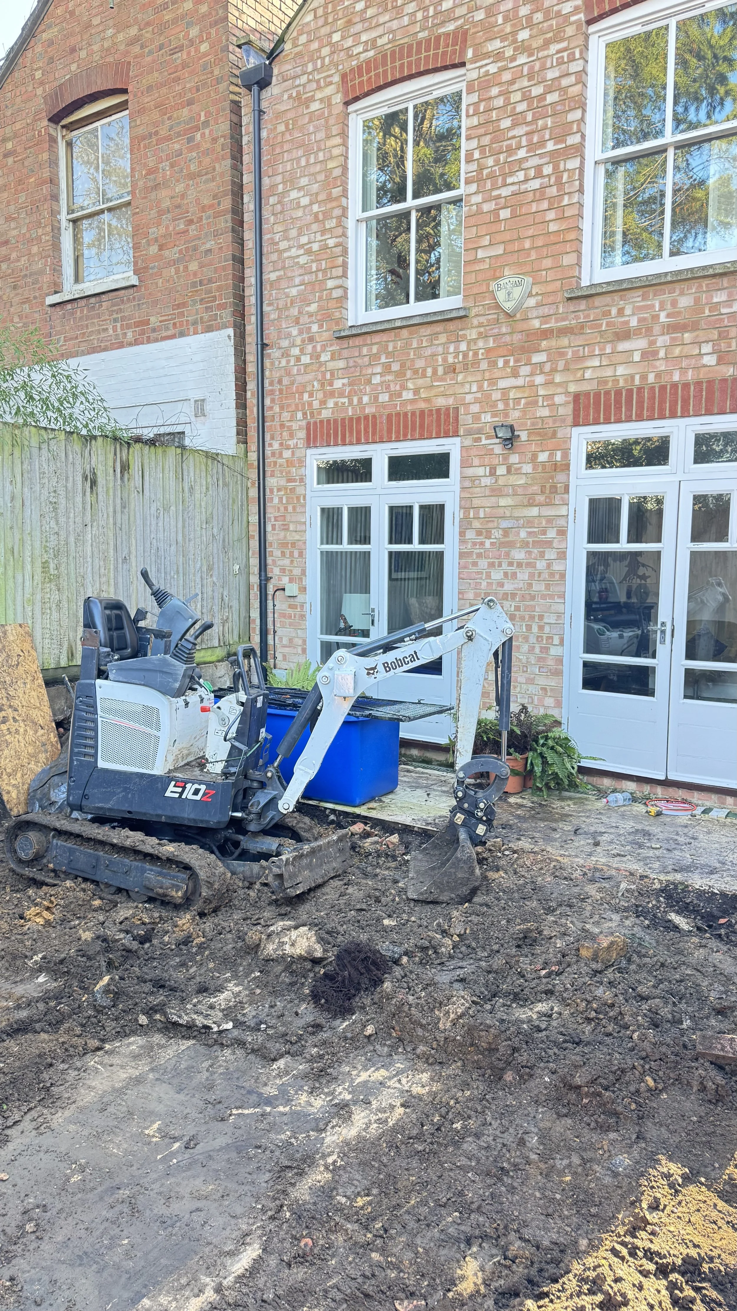 A small Bobcat E10z excavator parked on a dirt backyard with a brick house in the background, featuring two large windows, patio doors, and a wooden fence.