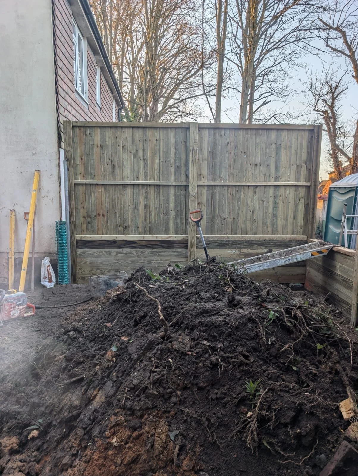 A backyard with a newly built wooden privacy fence, a mound of dirt and roots in the foreground, a snow shovel and some gardening tools leaning against the house, and a portable toilet on the right.