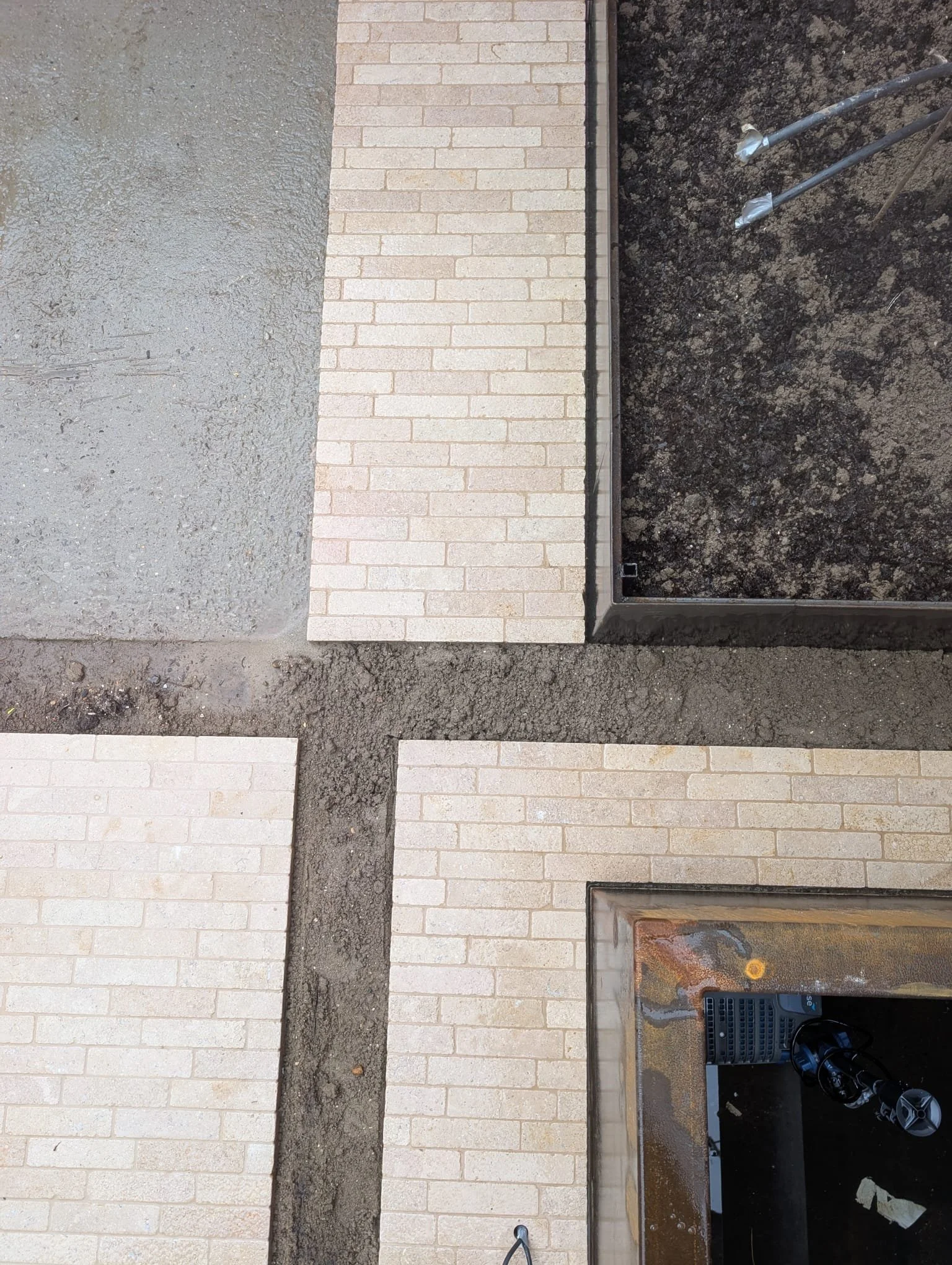 Exterior view of a building wall with beige bricks on the left and black bricks on the right, featuring a rectangular opening with a black metal grate, a water-damaged area at the bottom, and exposed metal pipes with plastic at the ends.