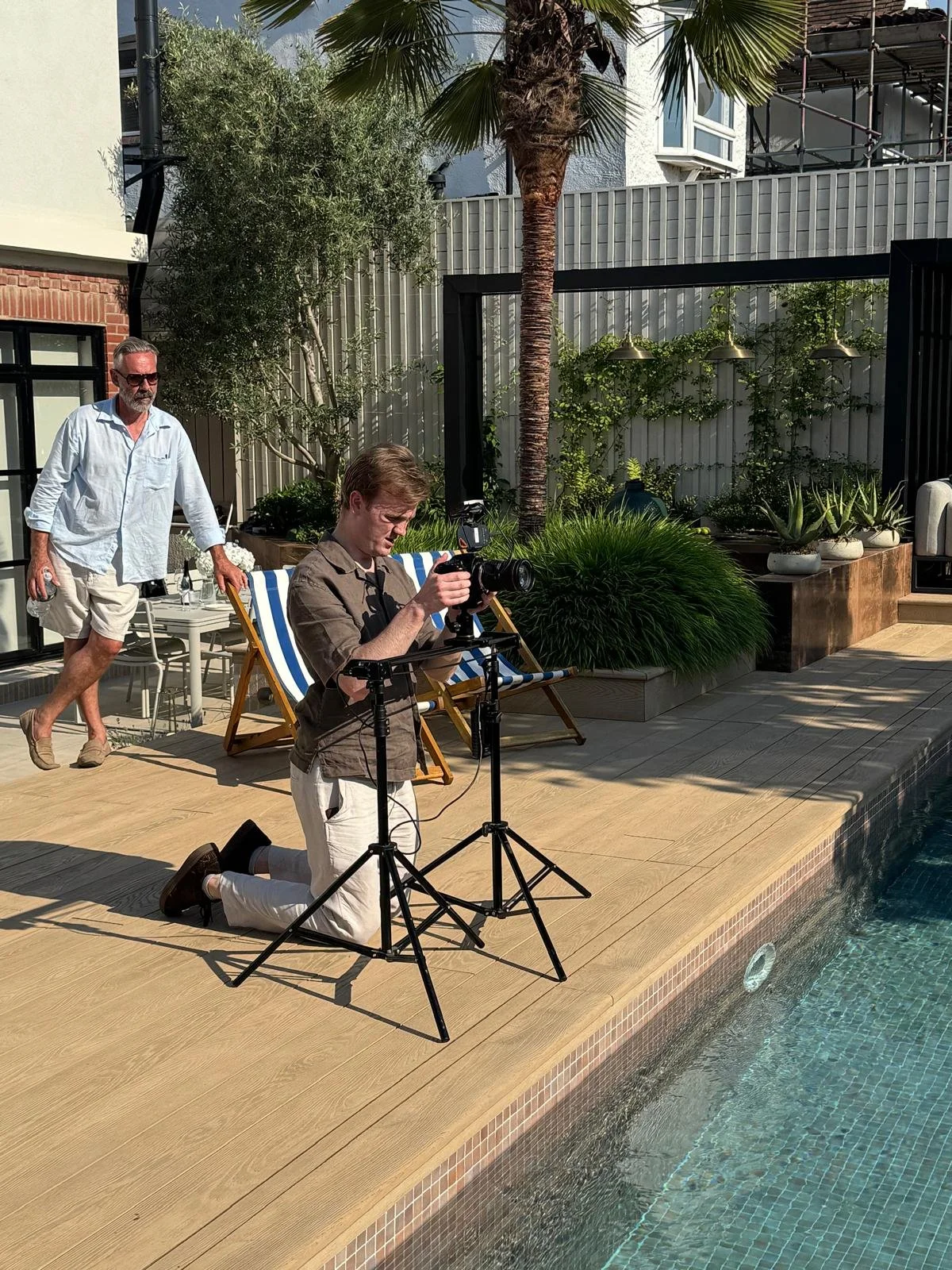 A person kneeling on a wooden pool deck, adjusting a camera on a tripod, near a swimming pool in a backyard with outdoor plants and furniture. Another person is walking in the background, wearing sunglasses, a light blue shirt, and shorts.