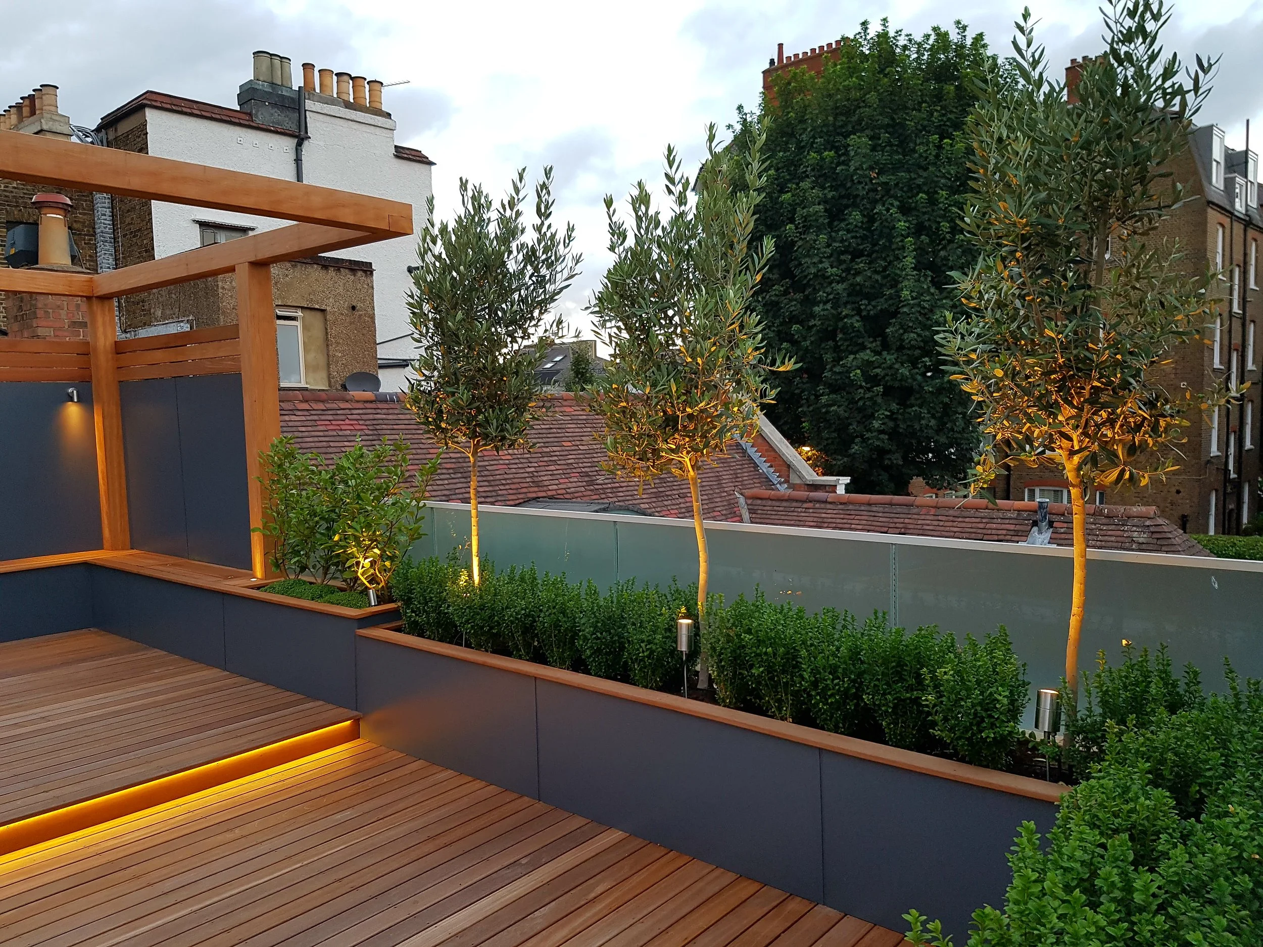 Urban rooftop patio with wooden decking, illuminated with yellow LED lighting, featuring potted trees and green bushes, surrounded by buildings and large trees in the background.