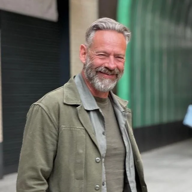 An older man with gray hair and a beard smiling, wearing a green jacket over a gray shirt, standing outdoors.