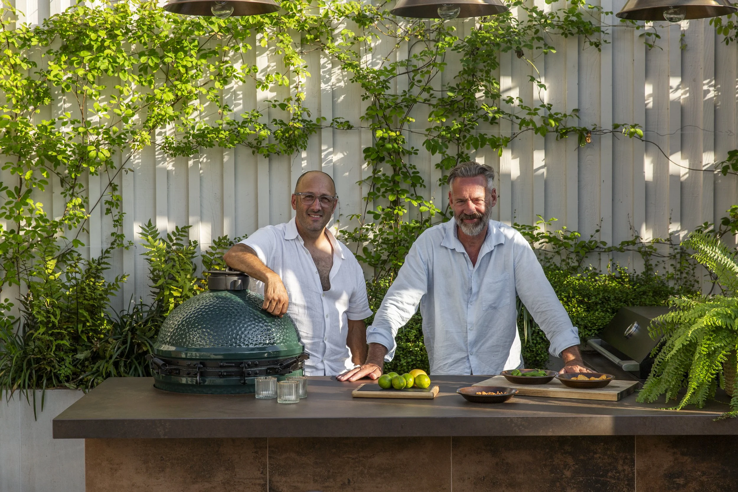 Two middle-aged men in white shirts standing behind a kitchen counter outdoors, surrounded by green plants and a white wooden fence. They are smiling, with grilling tools and food items like lime, plate of cooked meat, and bowls on the counter. One m
