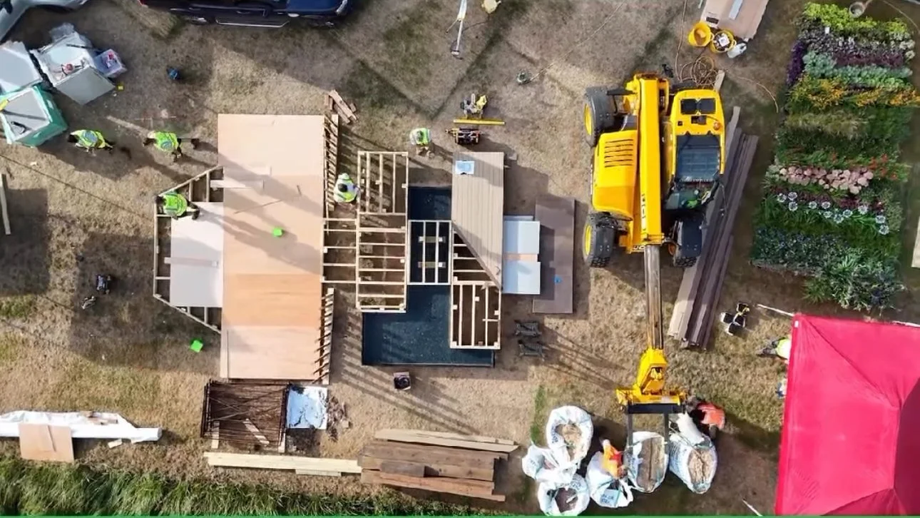 Aerial view of a construction site with workers building a garden, with a yellow construction vehicle and materials around, and a garden with flowers nearby.