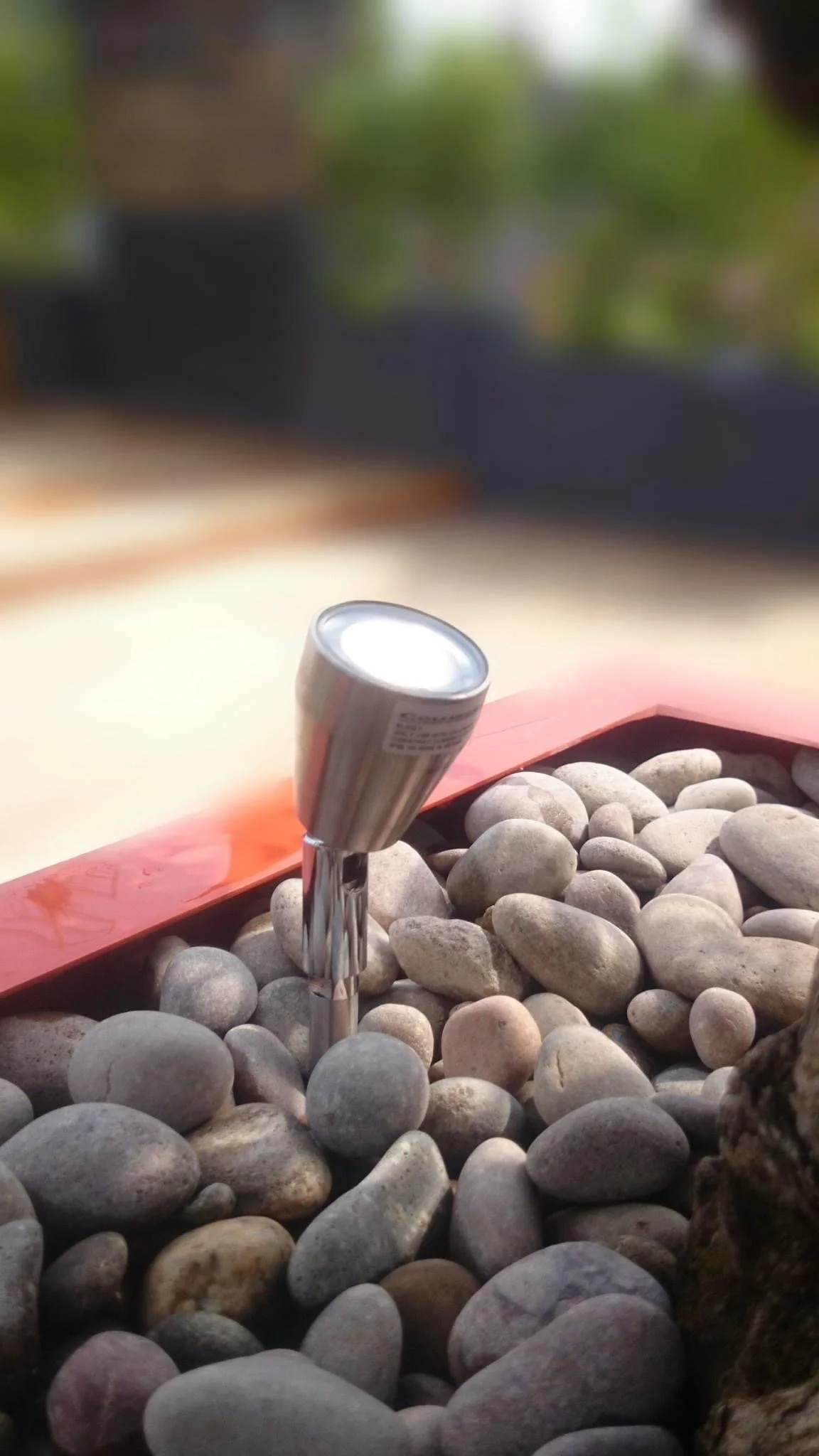 Close-up of a small outdoor water fountain with stainless steel spout among smooth round gray and beige stones, with blurred background of greenery