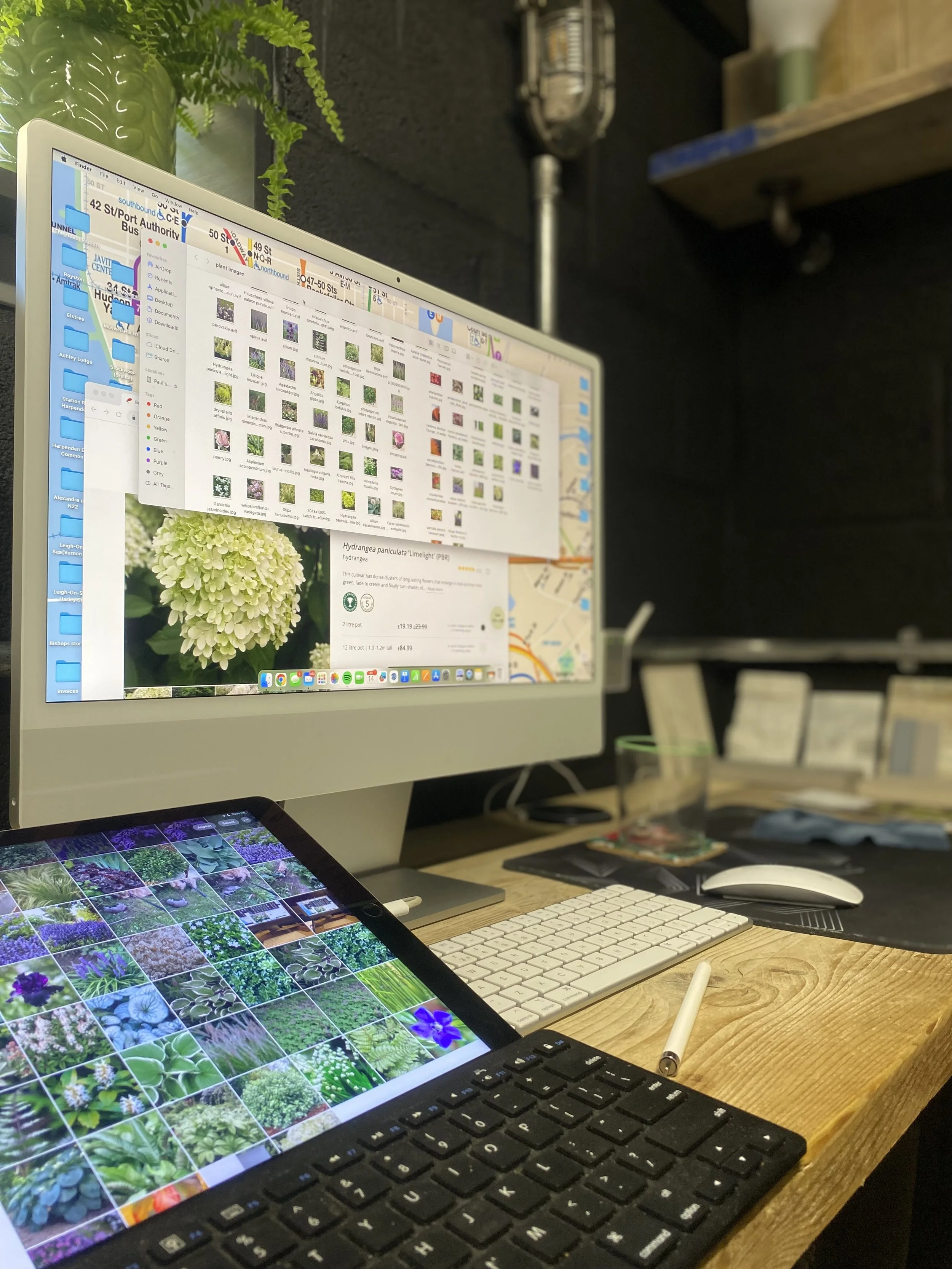 Desk workspace with a computer monitor displaying a photo library, a tablet showing various flower images, a wireless keyboard, a white stylus, a mouse, a glass with a lid, and a small green plant in a container. The workspace has a rustic wooden des