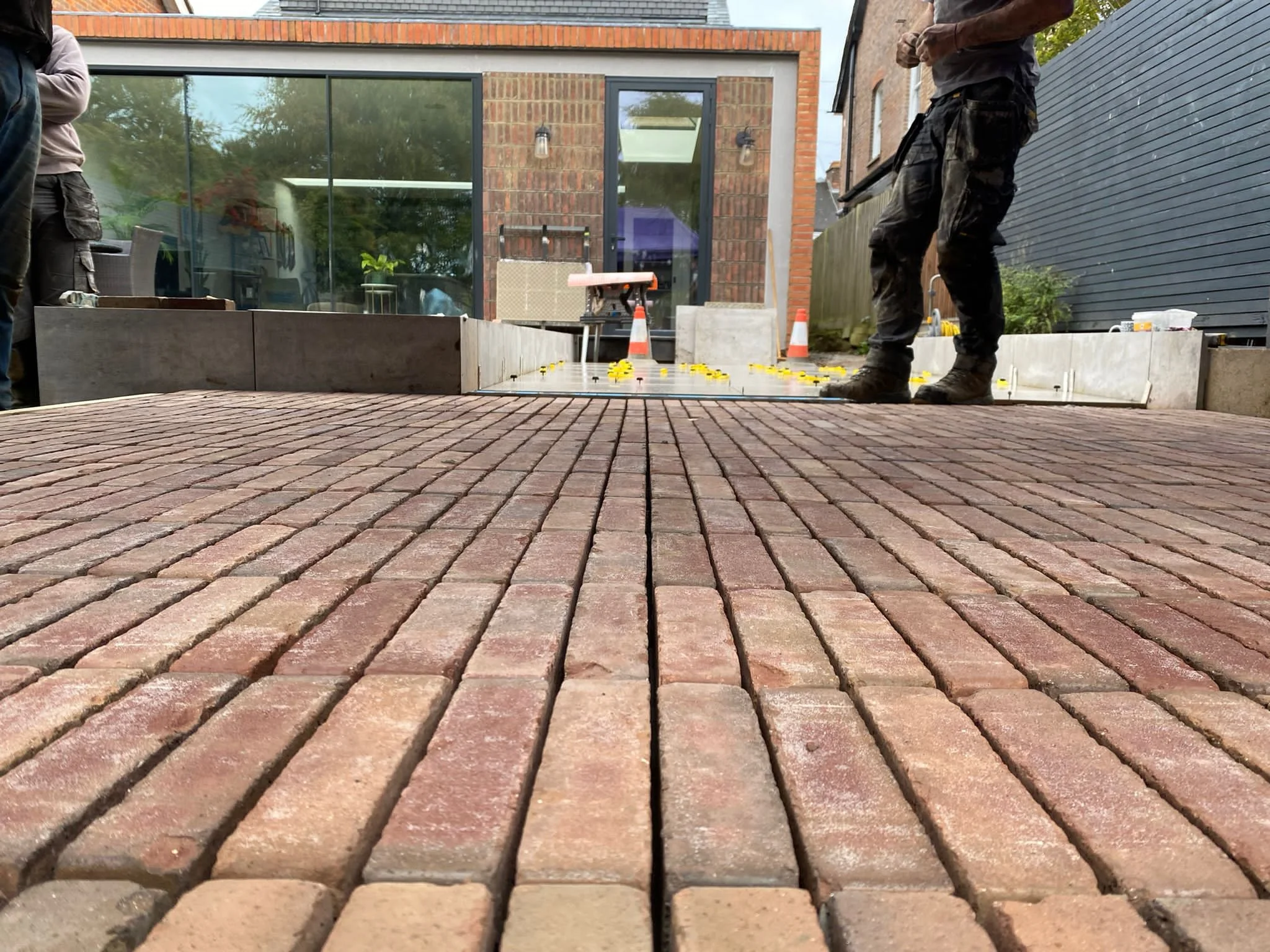 Close-up of a brick patio being laid by construction workers, with one worker standing on the bricks and tools and traffic cones in the background.