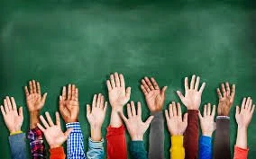 Multiple students raising their hands in a classroom with a green chalkboard in the background.