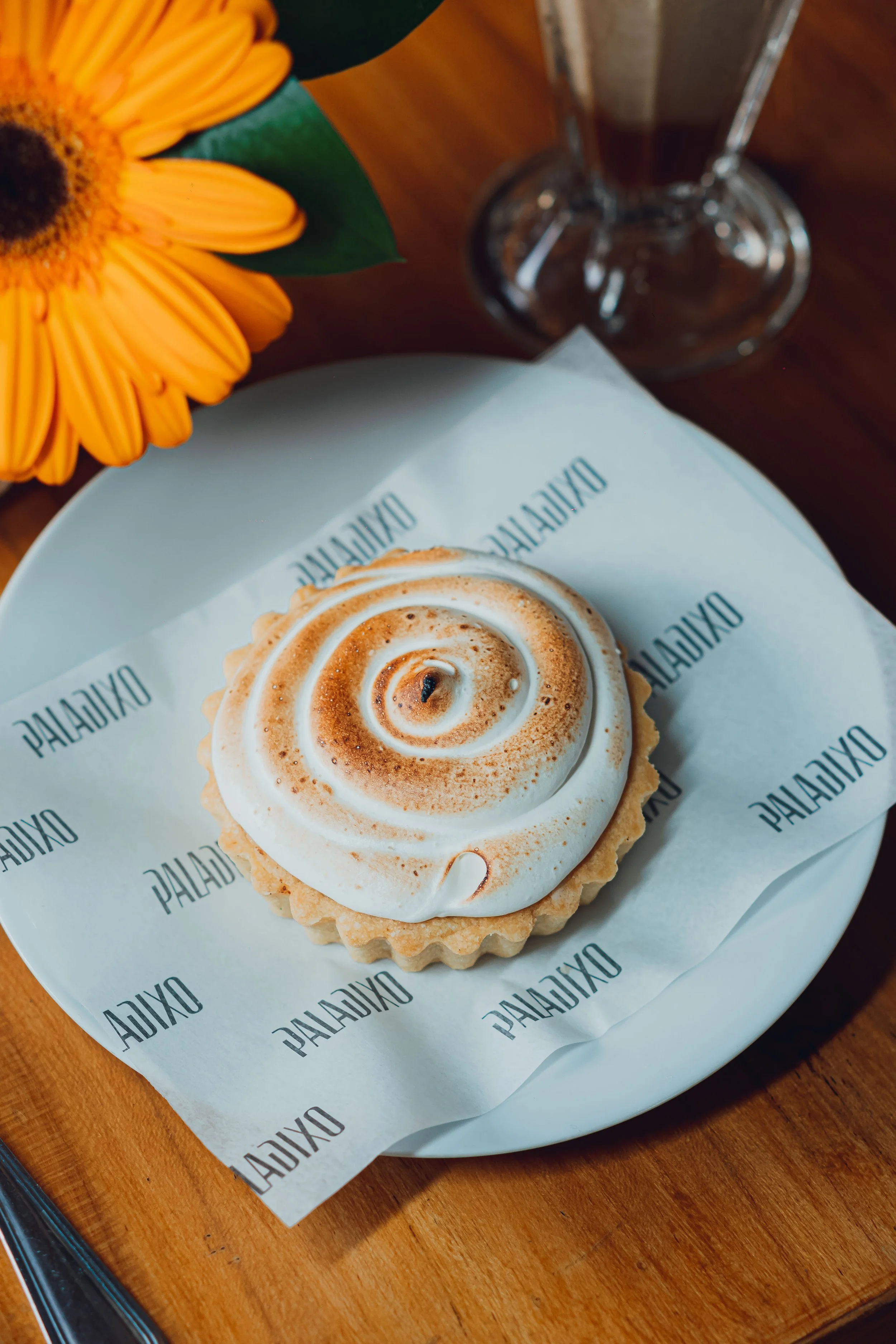 A lemon tart with meringue topping, served on branded paper on a white plate, with a yellow sunflower flower and a glass of dark beverage on a wooden table.