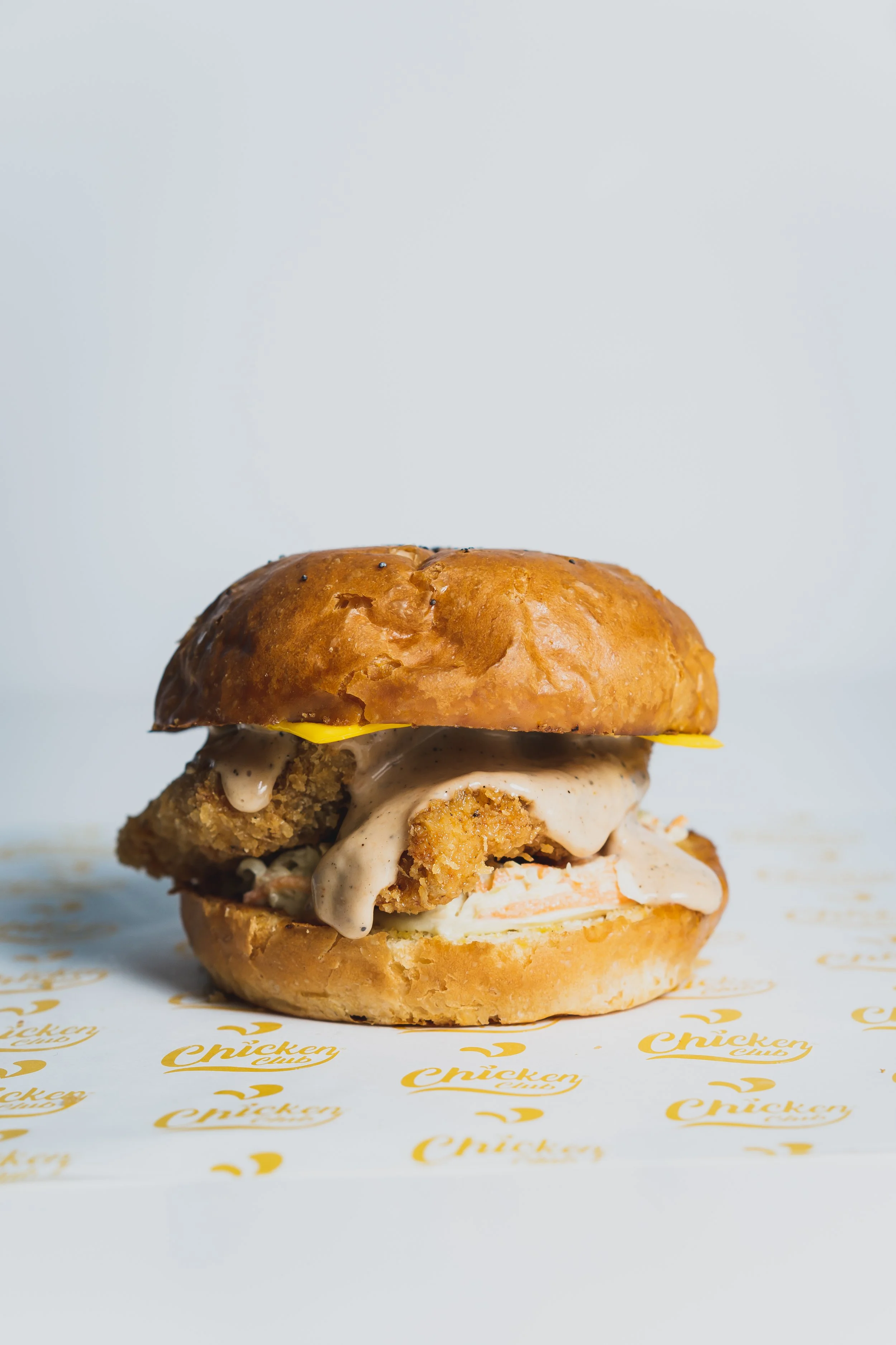 Close-up of a fried chicken sandwich with sauce, pickles, and melted cheese inside a soft bun on a branded paper.