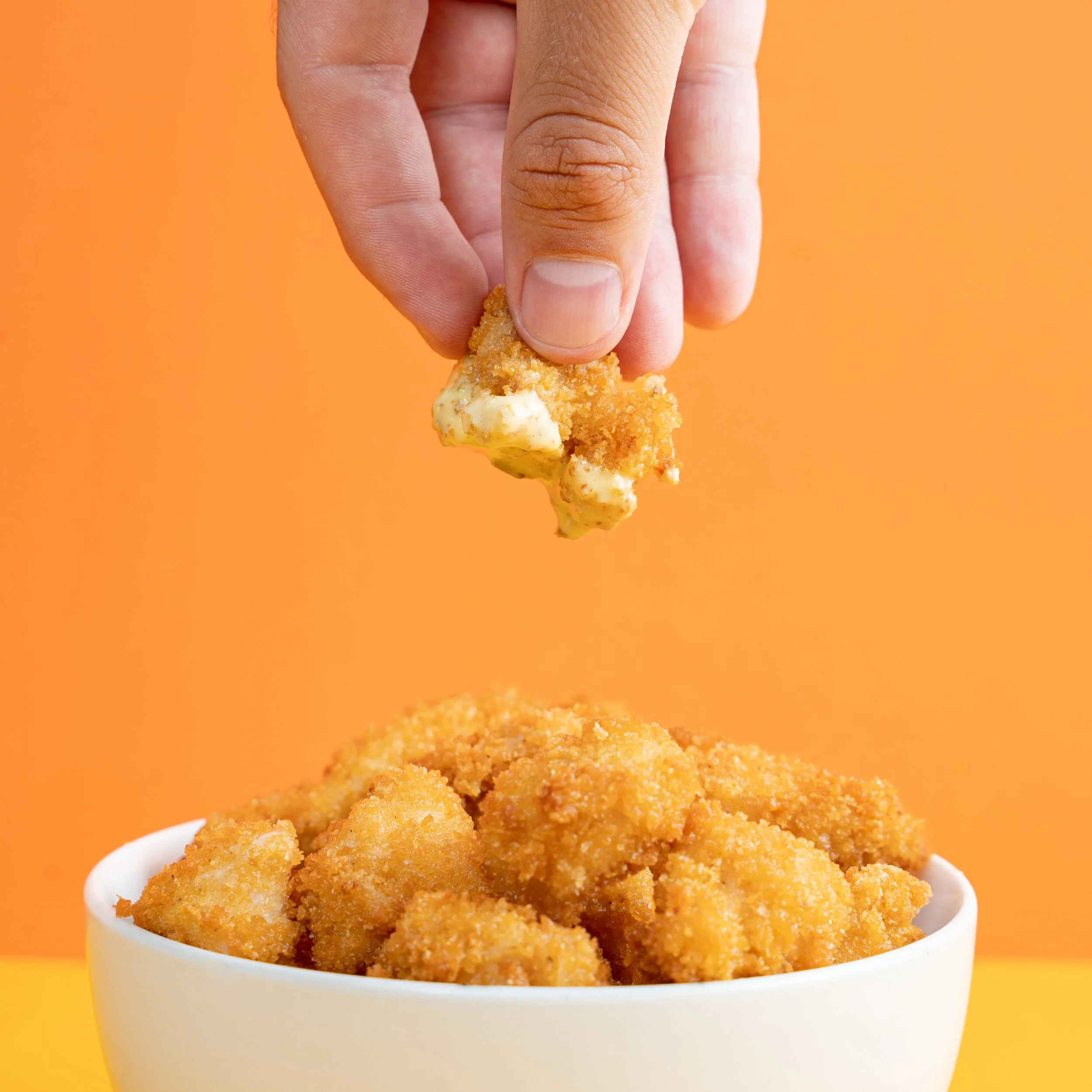 Persona tomando un trozo de pollo frito sobre un cuenco lleno de pollo frito en fondo naranja.