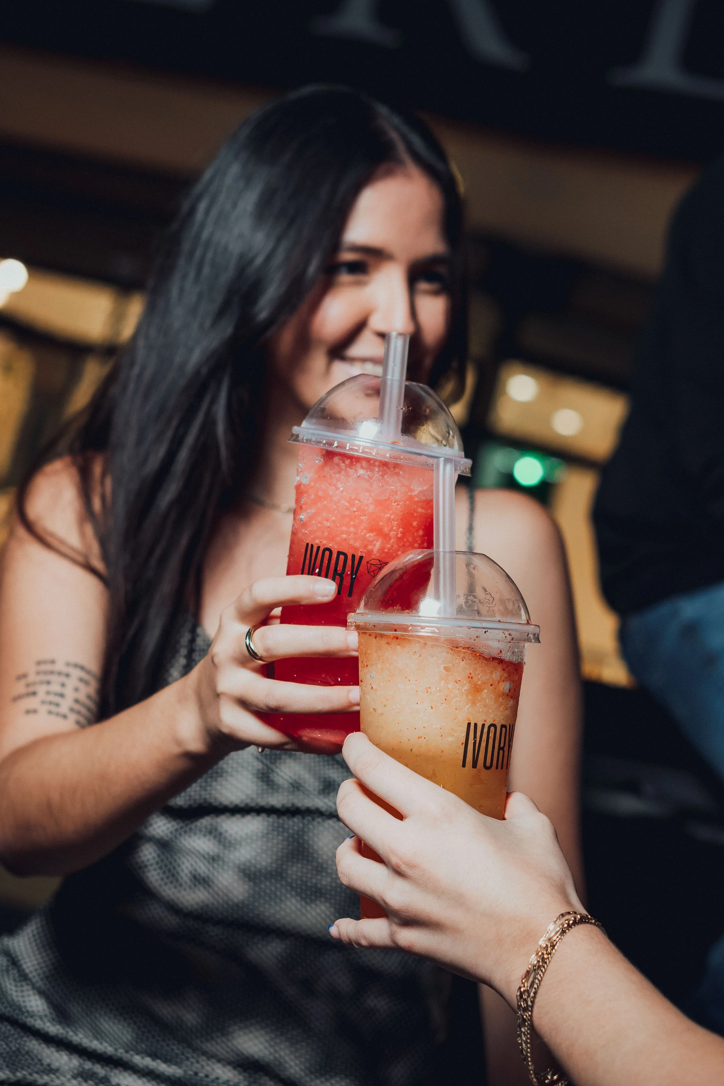 Two women holding colorful drinks in plastic cups with straws, smiling at each other in a social setting.