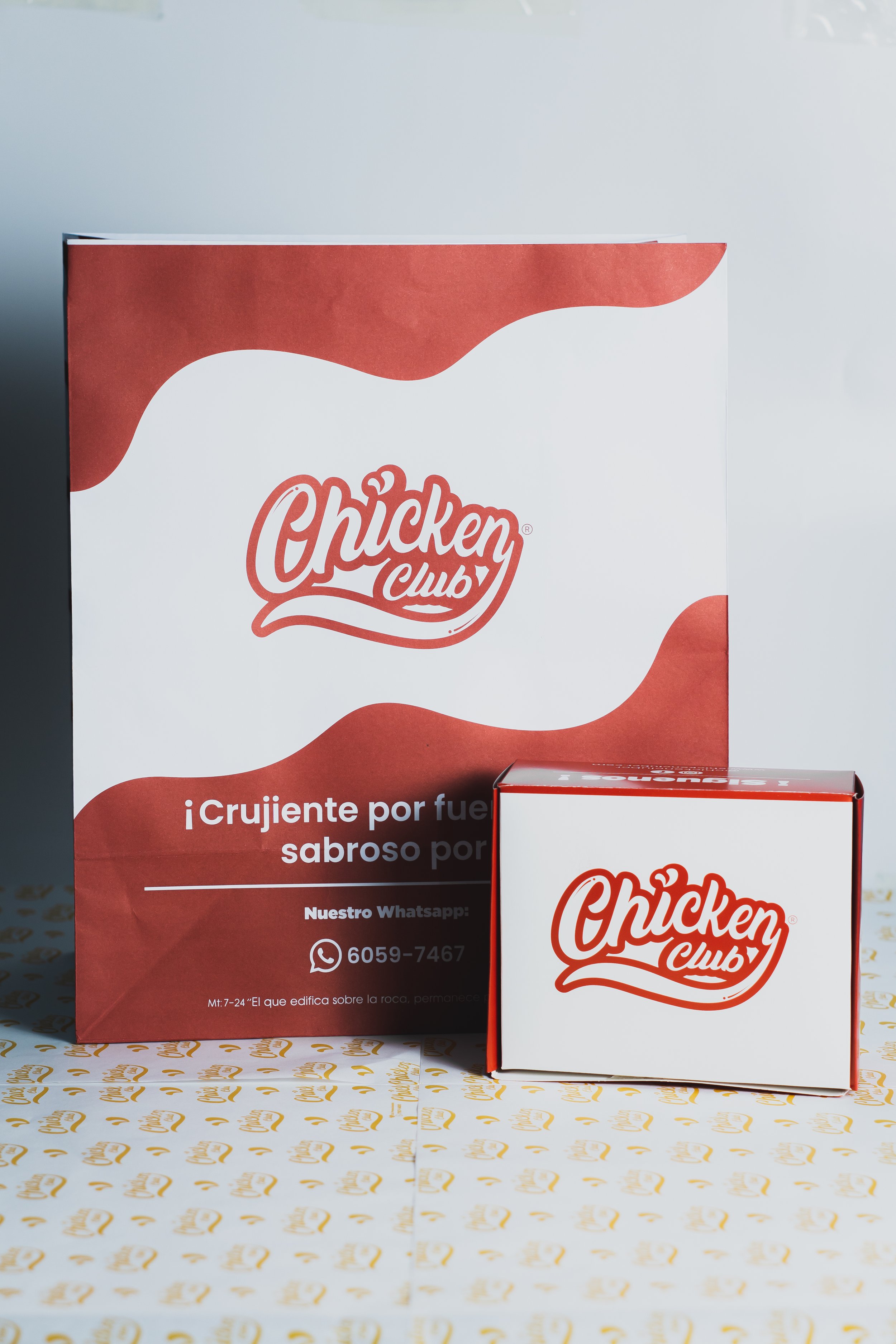 A white box with red accents and the logo 'Chicken Club' in red text, positioned next to a larger card with the same logo and text in Spanish, on a table with fast-food branding paper.